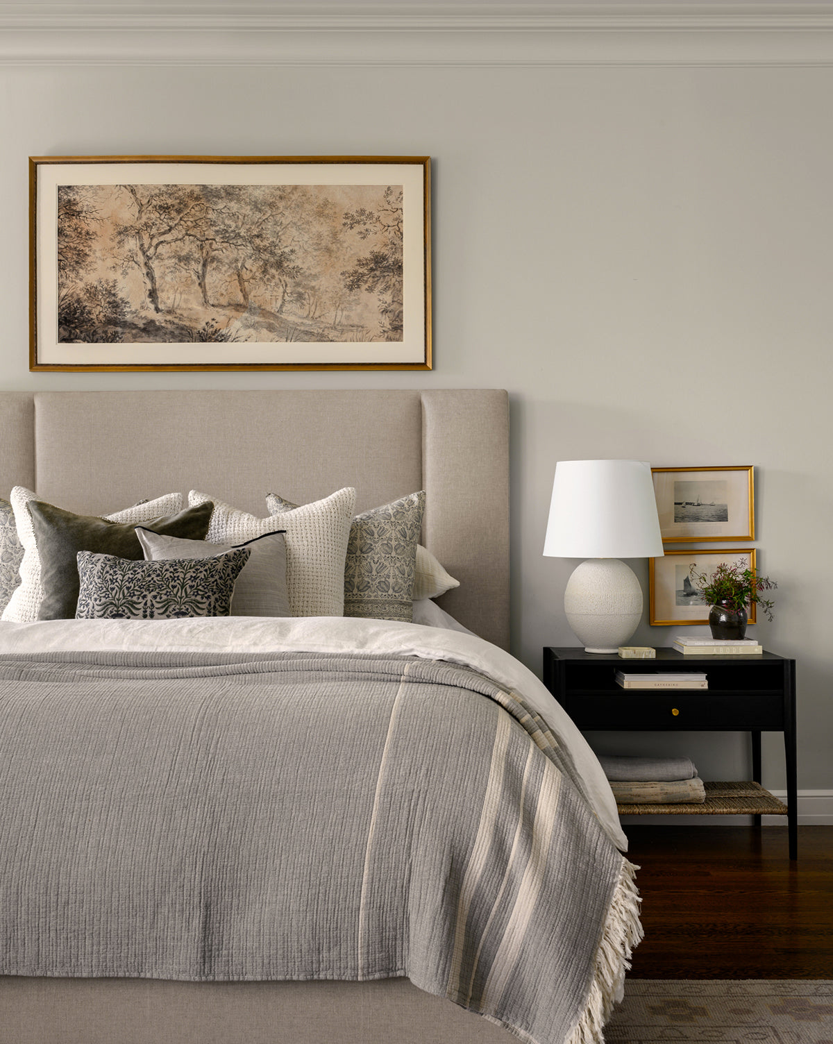 A neatly made bed with a beige upholstered headboard features throw pillows and a gray blanket. On the black nightstand, the Toulon Table Lamp with a linen shade sits alongside books, framed art, and flowers. A framed landscape hangs above the bed, by Vendixs