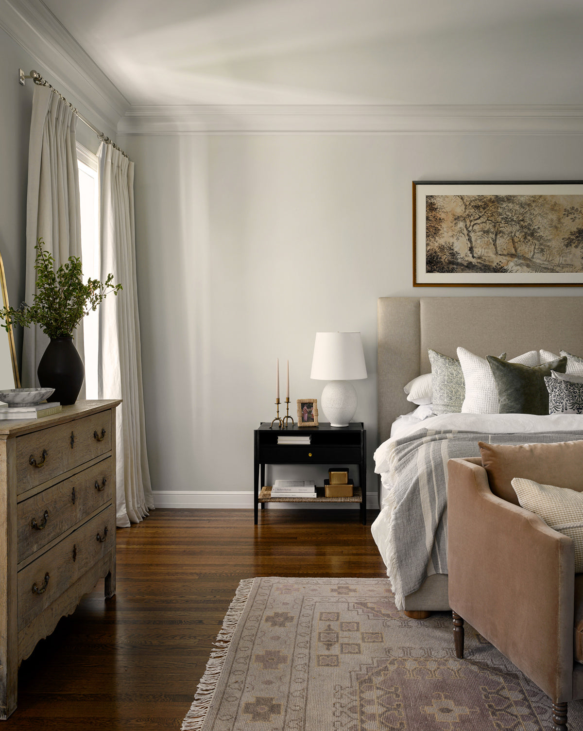 A cozy bedroom features a beige upholstered bed with layered pillows, a wooden nightstand and lamp, a vintage dresser with greenery, hardwood floors, and the Kenna Hand-Knotted Wool Rug—a perfect complement to the large landscape painting, by Vendixs