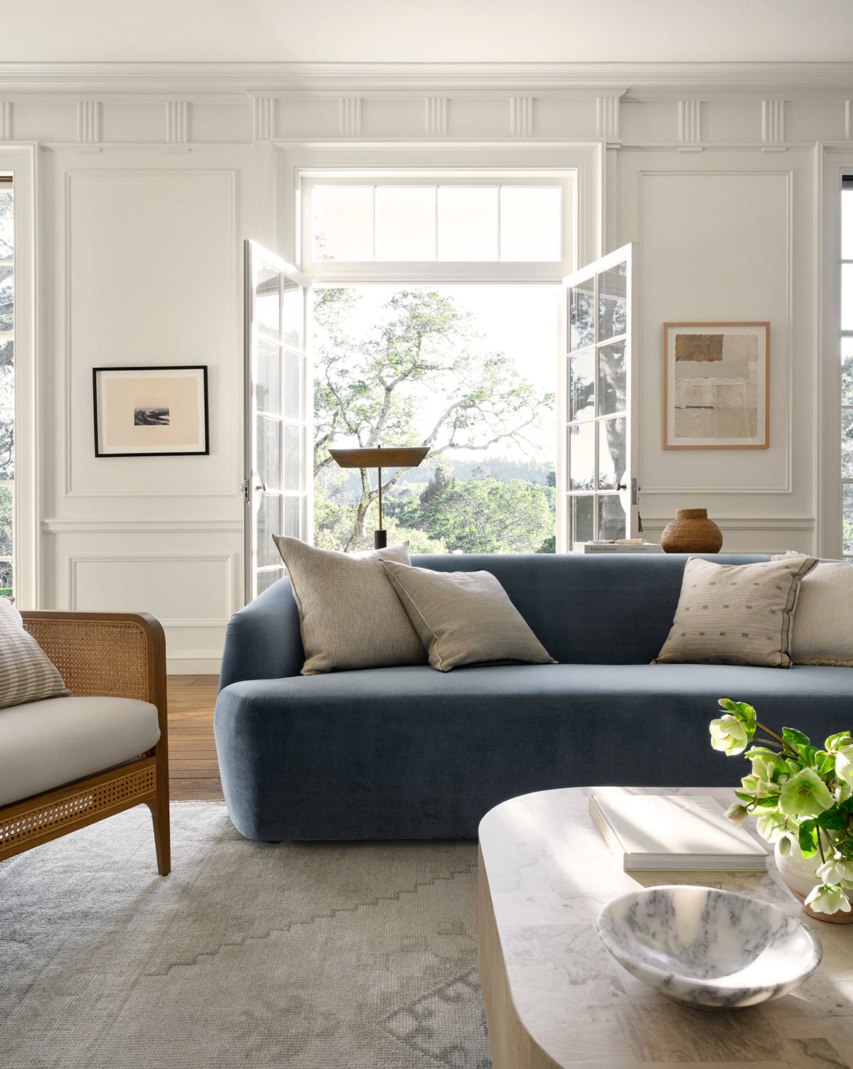 A bright, elegant living room features the Makerspalm Byron Sofa 96 with light cushions, a wooden armchair, art on white walls, large French doors opening to greenery, and a coffee table with a bowl, book, and flowers—styled by Vendixs