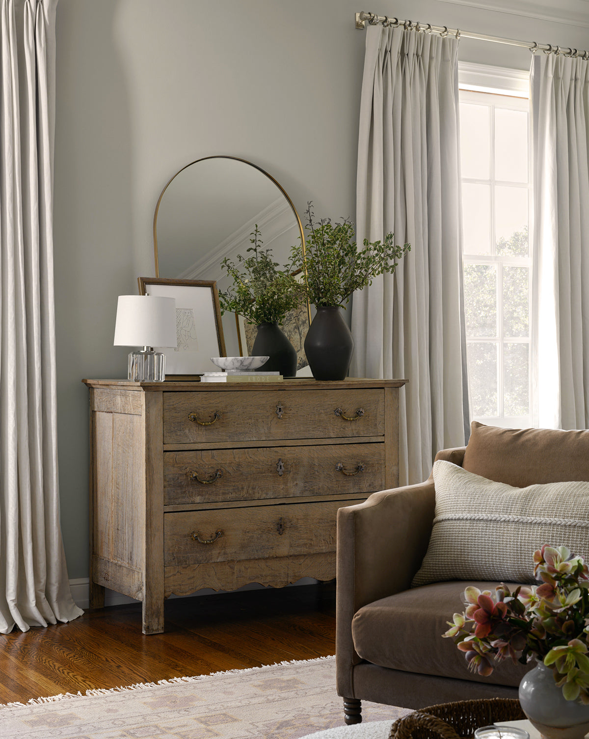 A cozy living room corner with a brown sofa, a wooden dresser topped with greenery vases, the Wylie Arched Mirror featuring shatterproof glass, framed art, and a white lamp. Light curtains frame a sunlit window for an inviting feel, by Vendixs