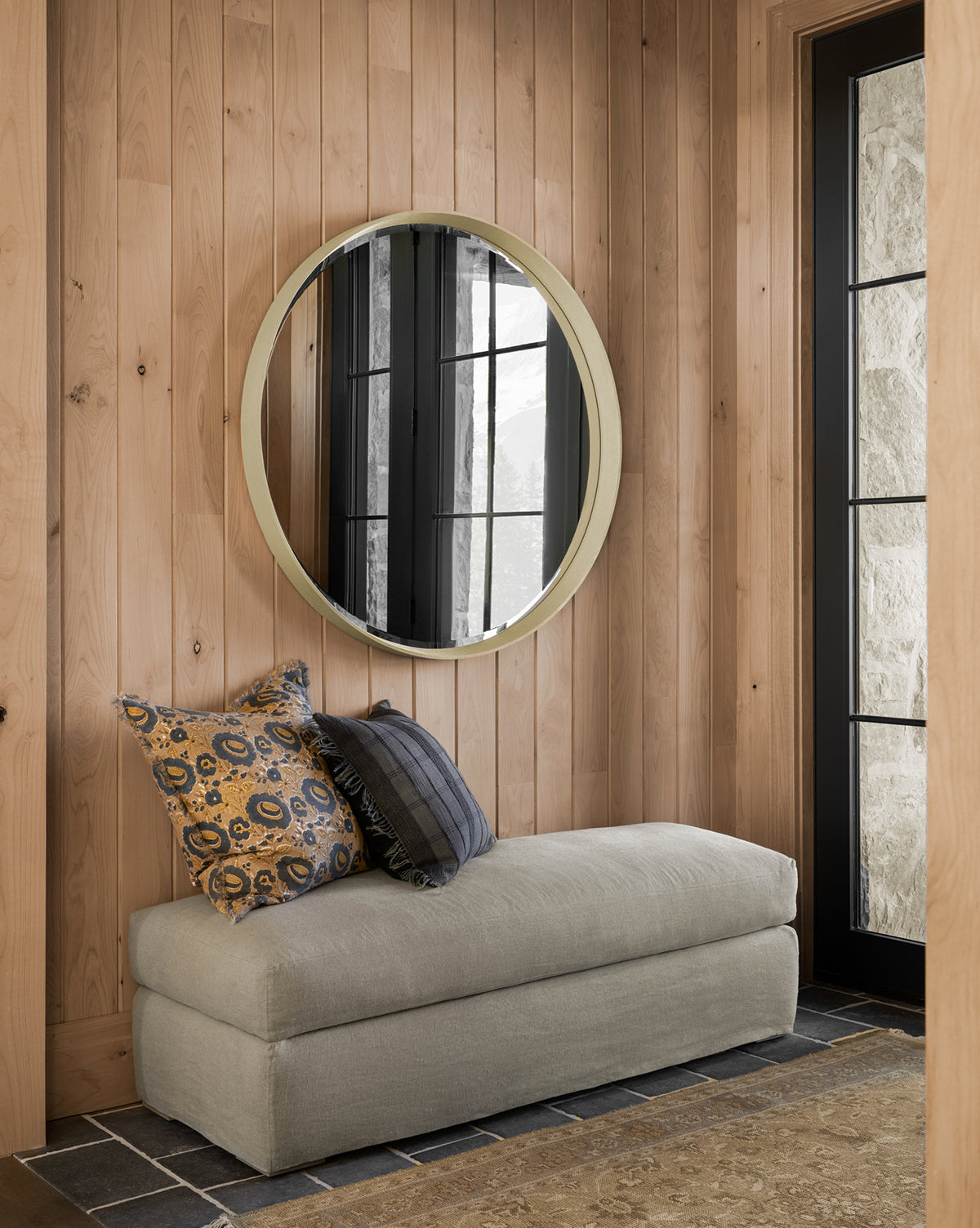 A cozy corner with light wood paneling, a round mirror, beige bench, and patterned pillows. Black-framed windows brighten the space, showcasing stylish living room furniture and the customizable Macy Slipcover Ottoman by Community—by Vendixs