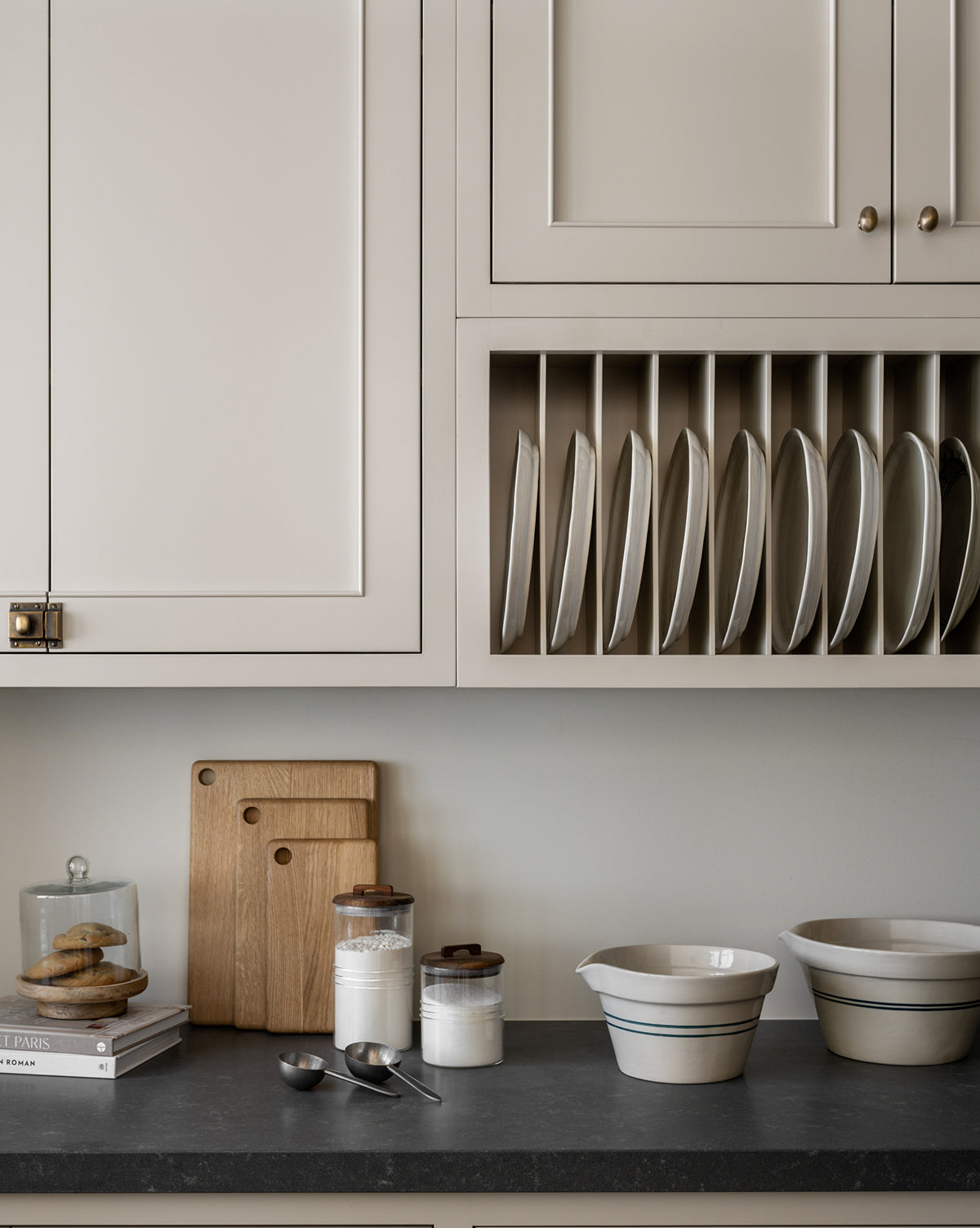 A modern kitchen counter with three wooden cutting boards, a glass jar of cookies, a stack of books, jars of flour and sugar, and an Everett Mixing Bowl, by Vendixs Plates are neatly stored vertically in a rack above the counter.
