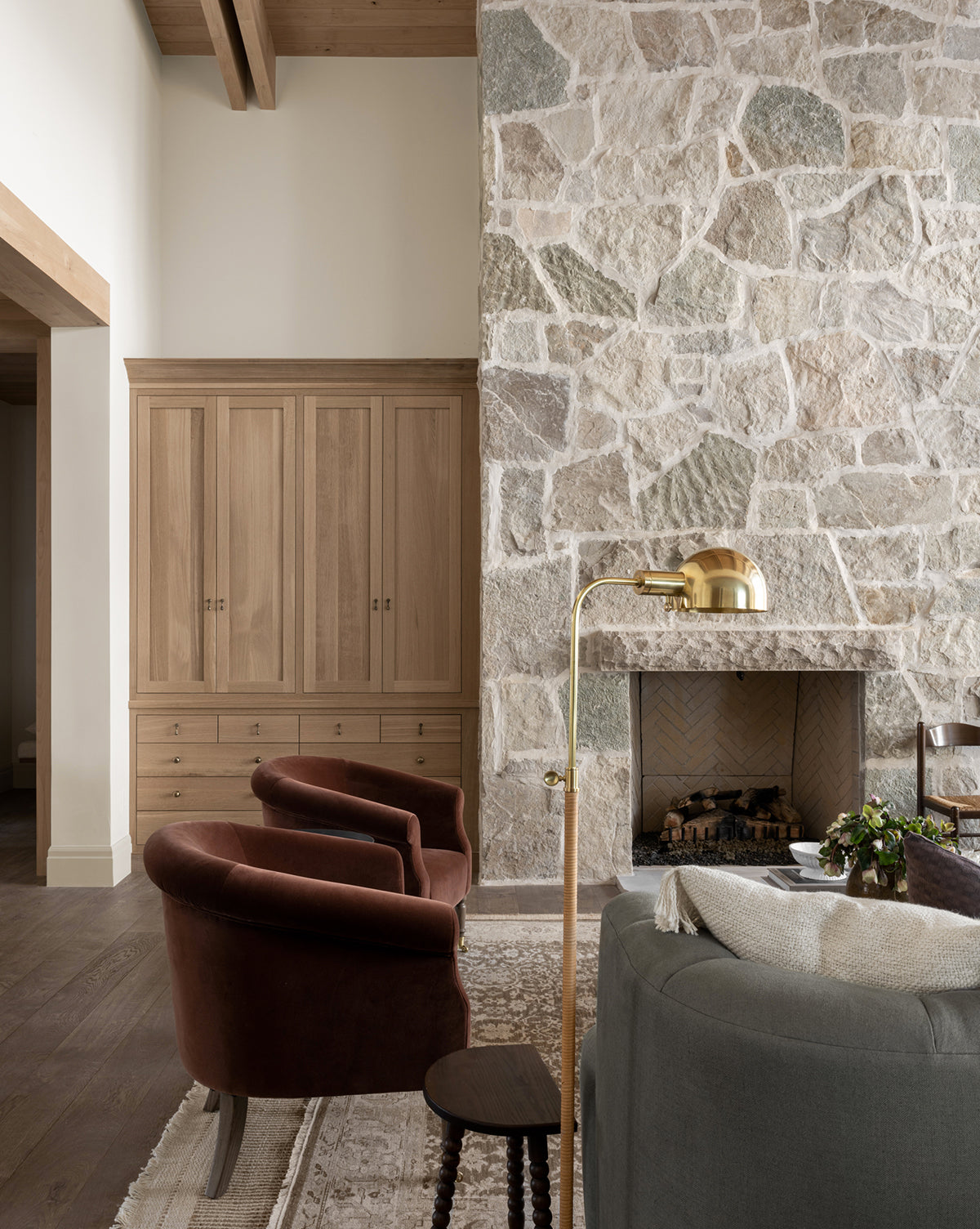 A cozy living room with two brown velvet chairs, a gray sofa, stone fireplace, wooden cabinet, and the Devon Floor Lamp (aged bronze with rattan base), all set on a patterned rug over wooden floors and under exposed beams, by Vendixs