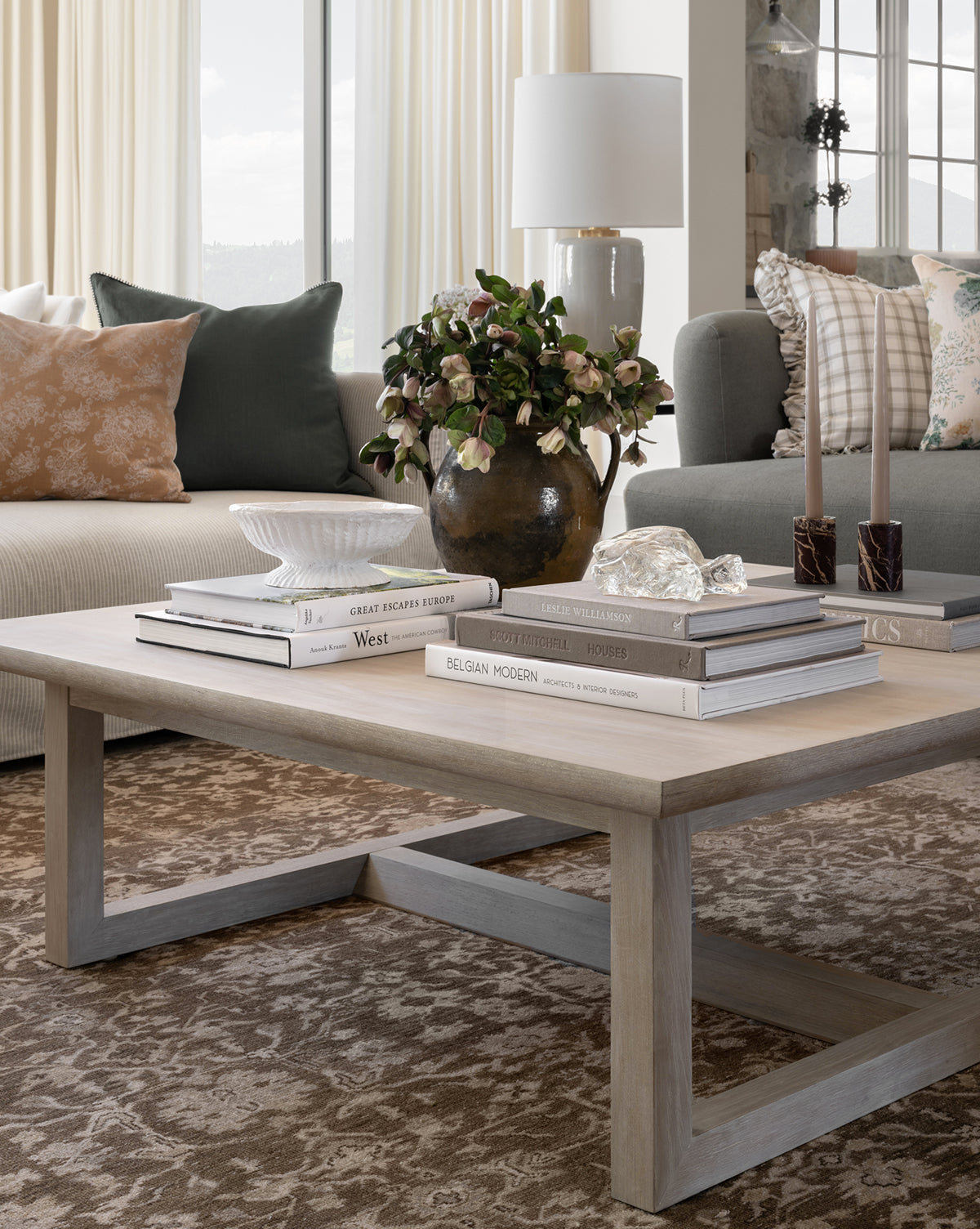 A modern living room features a beige sofa with patterned cushions and the Herrin Coffee Table displaying stacked books, a potted plant, a shell dish, glass decor, and candles on a brown patterned rug. Curtains and windows are in the background, by Vendixs