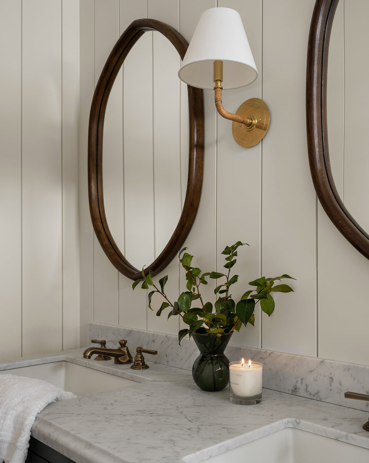 A bathroom vanity with a marble countertop, brass faucet, vase of green branches, lit candle, towel, oval Oslo Mirror with oak frame, and wall-mounted brass sconce with white shade, by Vendixs
