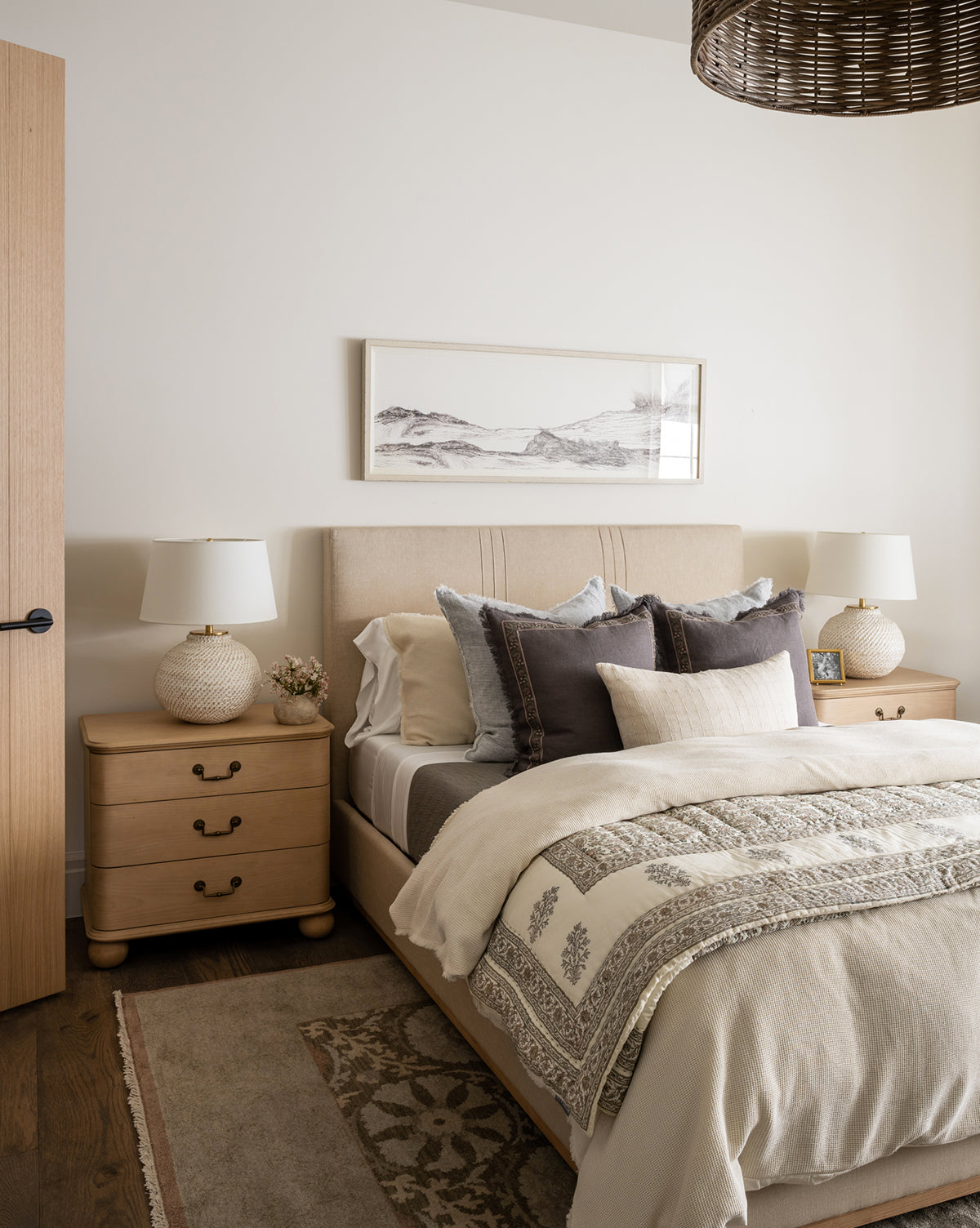 A cozy bedroom features a beige upholstered bed with neutral bedding, two wooden nightstands, and a Suzani Hand-Knotted Wool Rug with floral motifs. Framed landscape art hangs above the bed, and a woven light fixture completes the look, by McGee & Co.