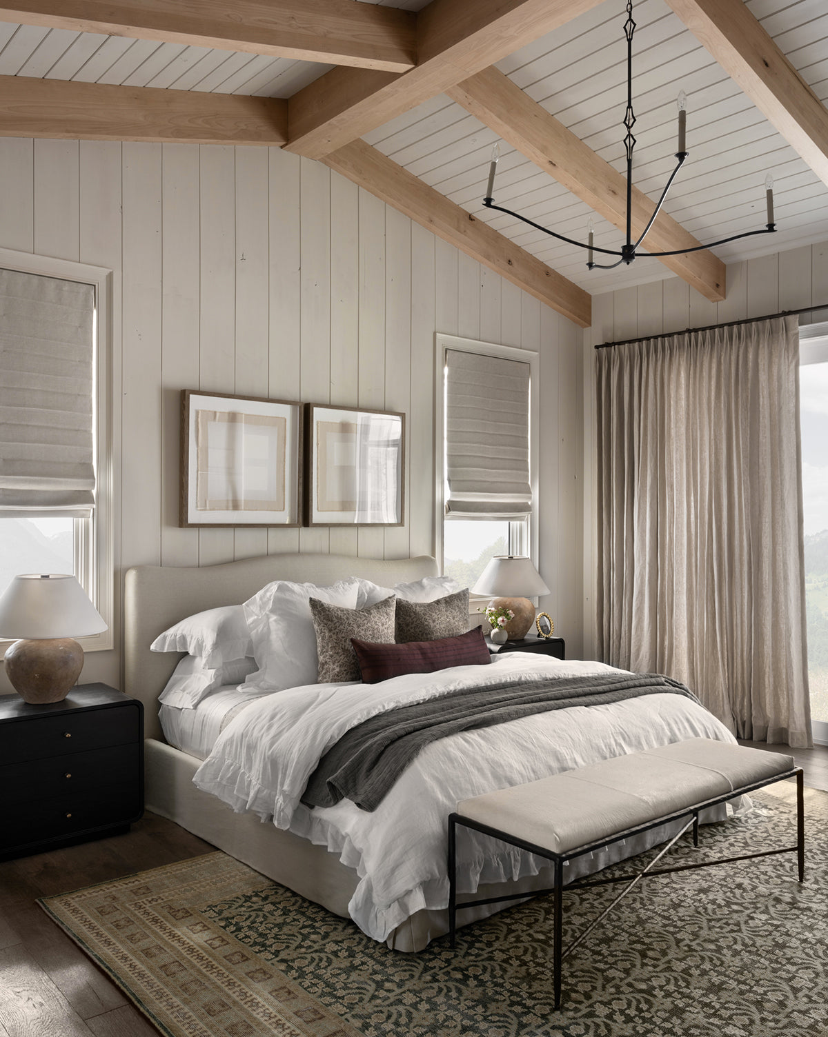 A cozy bedroom with a large bed, white layered bedding, decorative pillows, a foot bench, wood-beamed ceiling, side tables with lamps, two windows with roman shades, and the Evangeline Hand-Knotted Wool Rug for added texture, by Vendixs