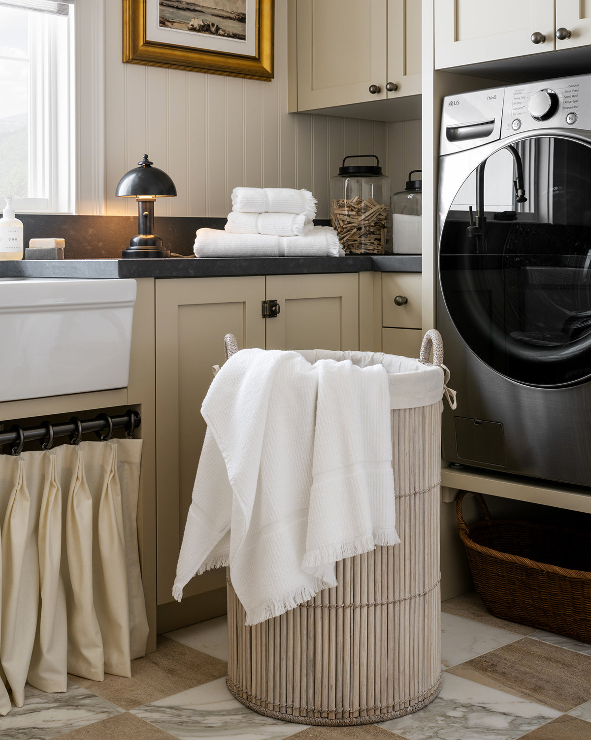 A tidy laundry room with a washer, folded towels, a wicker basket, and light cabinetry is brightened by the sleek Totie Task Lamp on the countertop with soap and storage jars, by Vendixs