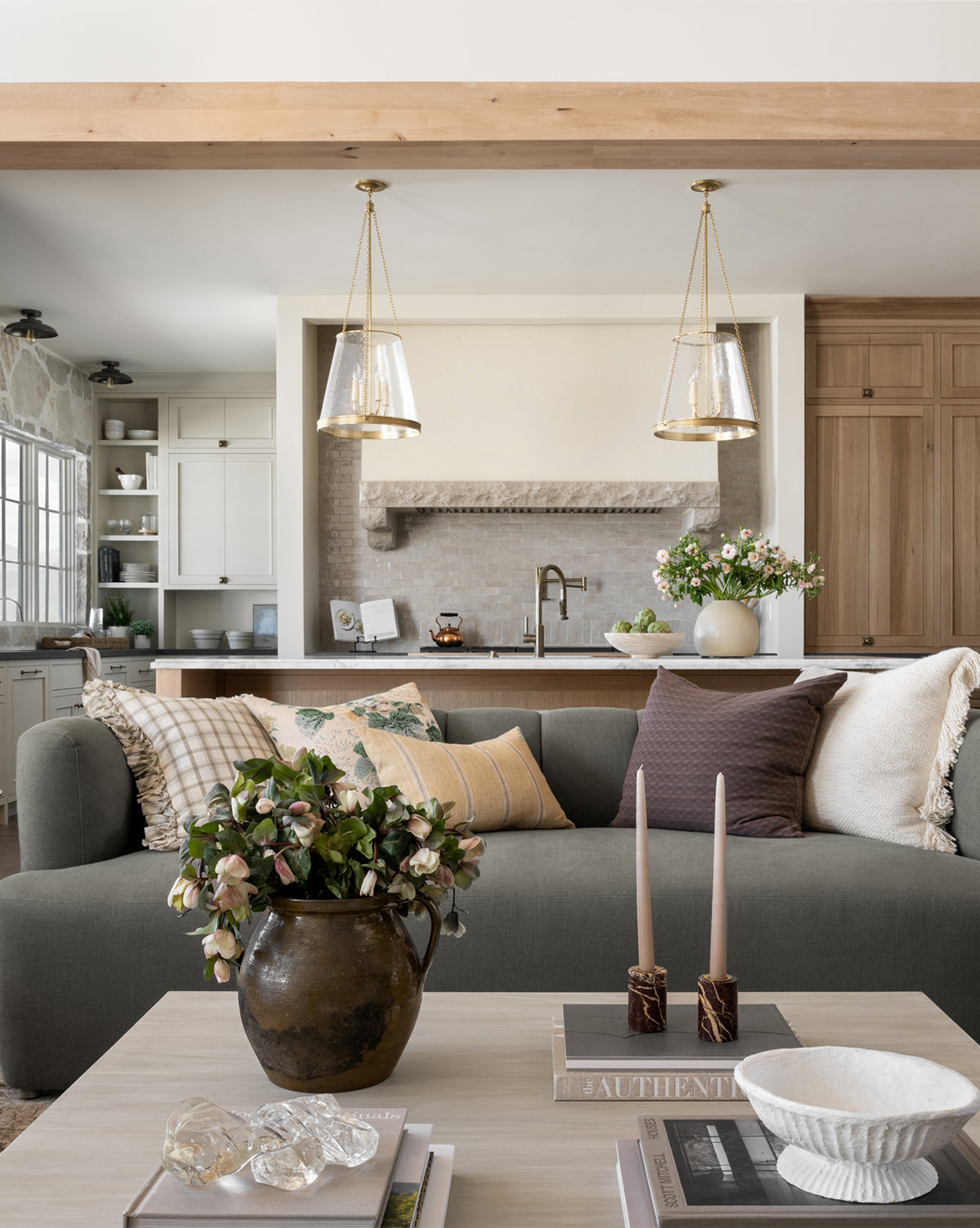 Stylish living room featuring a modern Ines Sofa 90 in gray with neutral pillows, plus a coffee table set with candles, a bowl, and a potted plant. The background highlights a contemporary kitchen with pendant lights and light wood cabinetry, by Vendixs