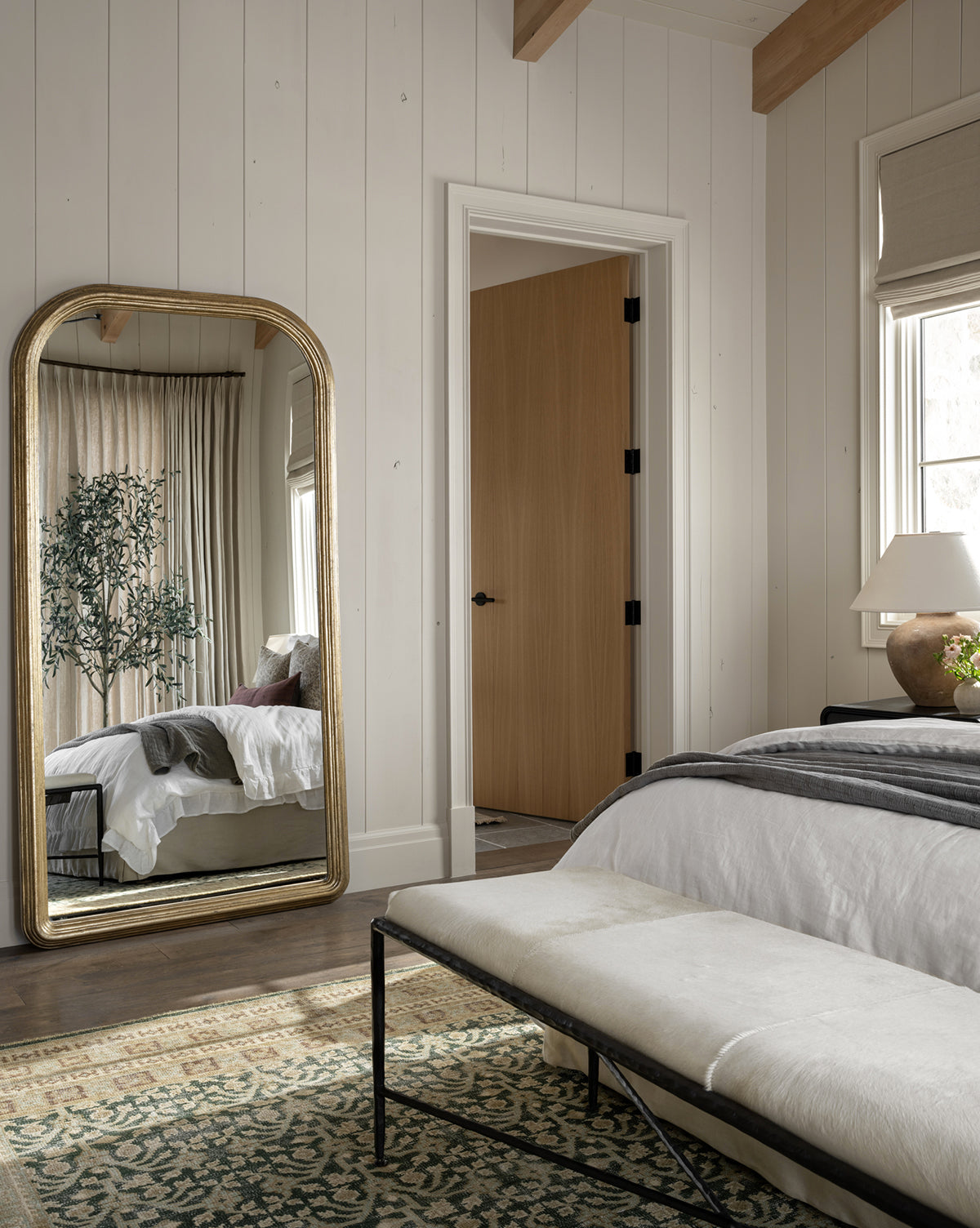 A cozy bedroom with light wood walls features the Giada Floor Mirror with an elegant frame, reflecting a bed with white and gray bedding, a potted plant, a bench at the foot of the bed, and natural light through beige curtains, by Vendixs