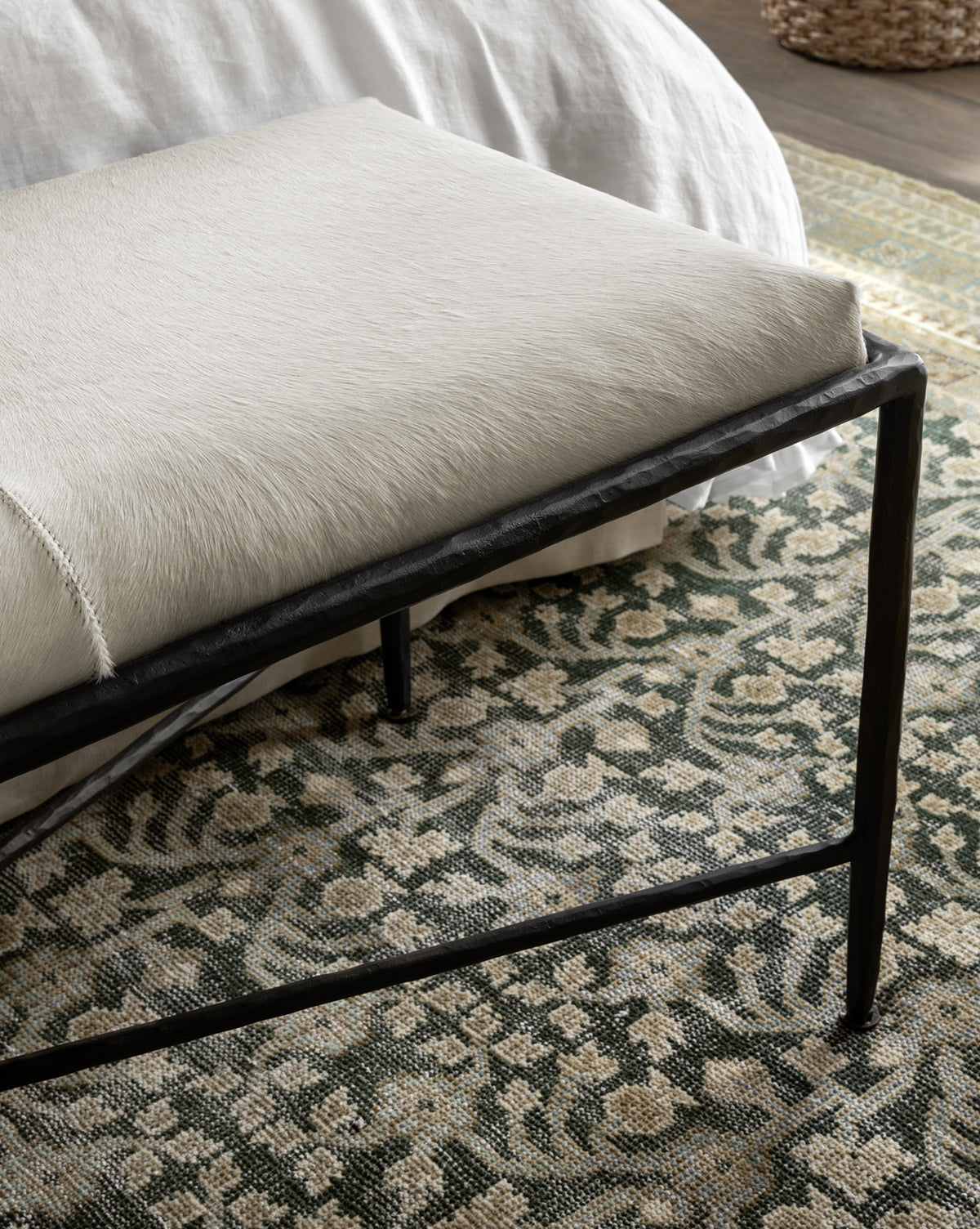 A close-up of a bench with a cream-colored upholstered seat and black metal legs, placed on the Evangeline Hand-Knotted Wool Rug with floral designs near a white bedspread, by McGee & Co.