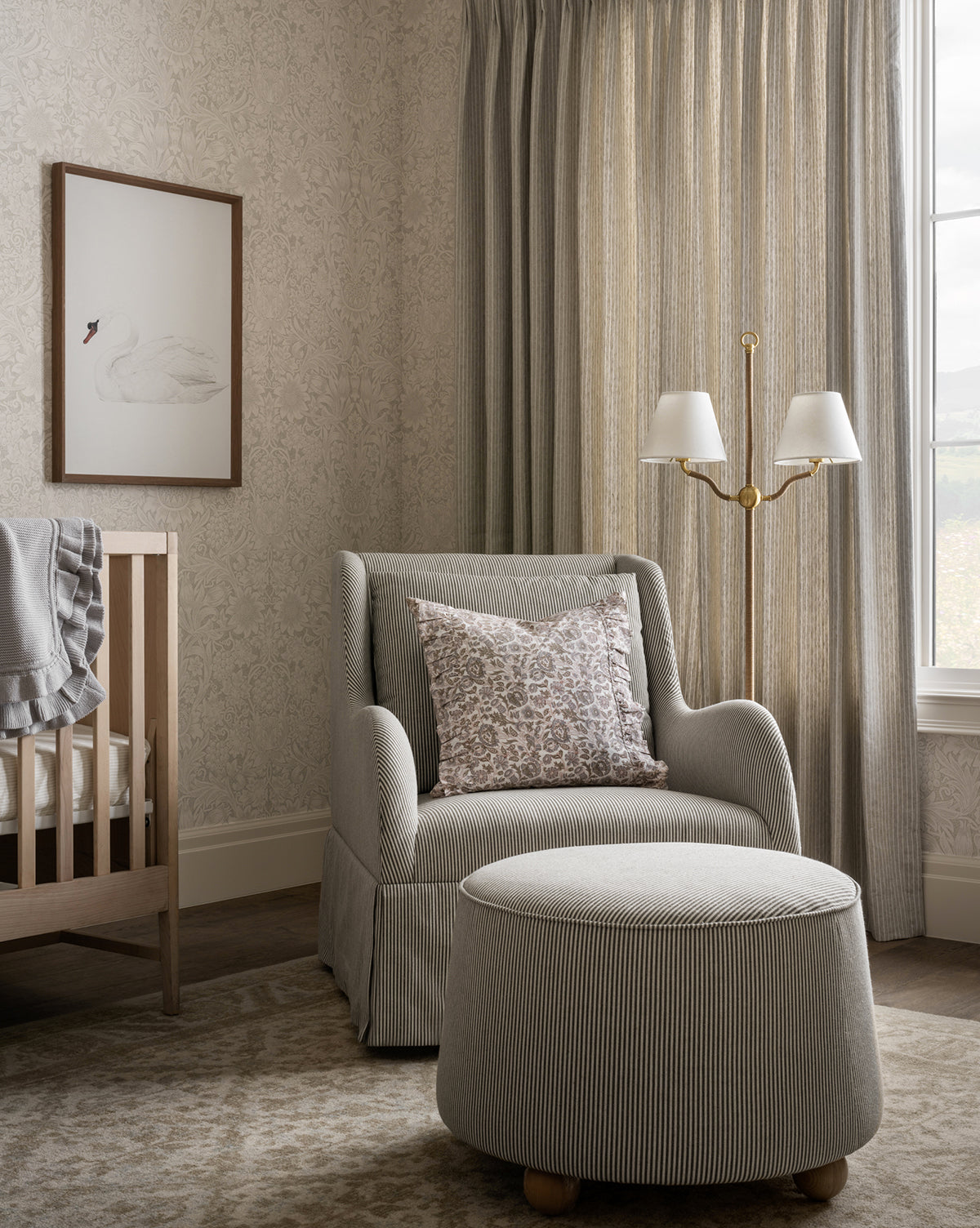 A cozy nursery with a crib, plush seat with the Barnaby Ottoman, floral pillow, dual-shade floor lamp, beige patterned curtains, framed swan art, and softly patterned wallpaper by Vendixs
