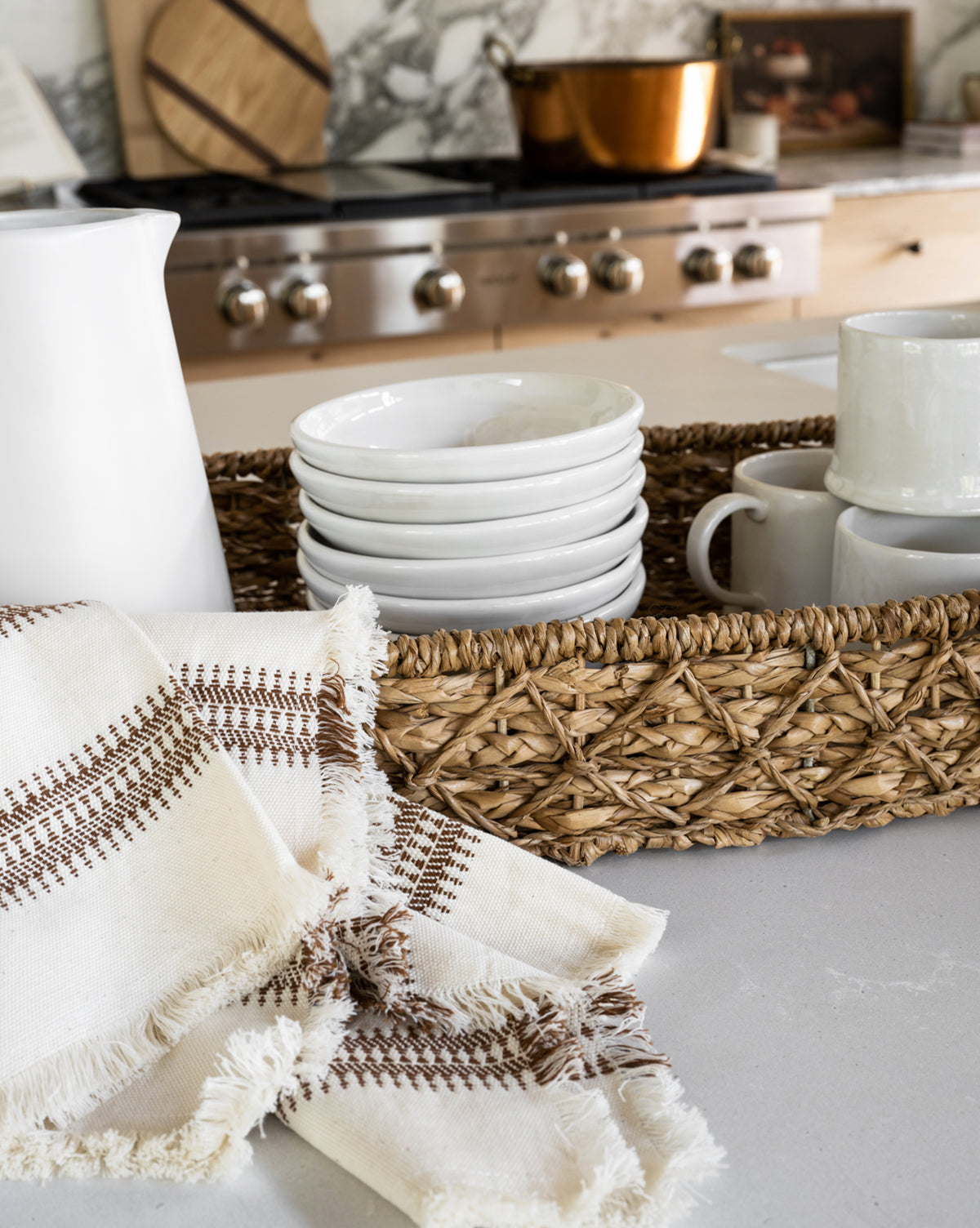 A woven basket holds stacked white bowls and mugs on a kitchen counter. In the foreground, a Woven Stripe Tea Towel with brown stitching and fringe is displayed, with a stove and copper pot visible in the background, by Vendixs