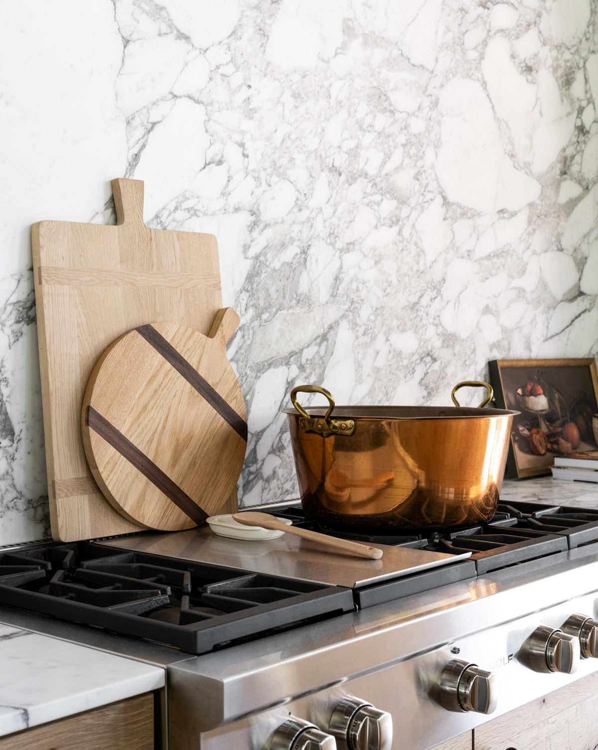 A kitchen stove with a large copper pot, a wooden spoon, two cutting boards—one being the Round Oak Bread Board—and a small framed artwork resting against a white and gray marble backsplash, by Vendixs
