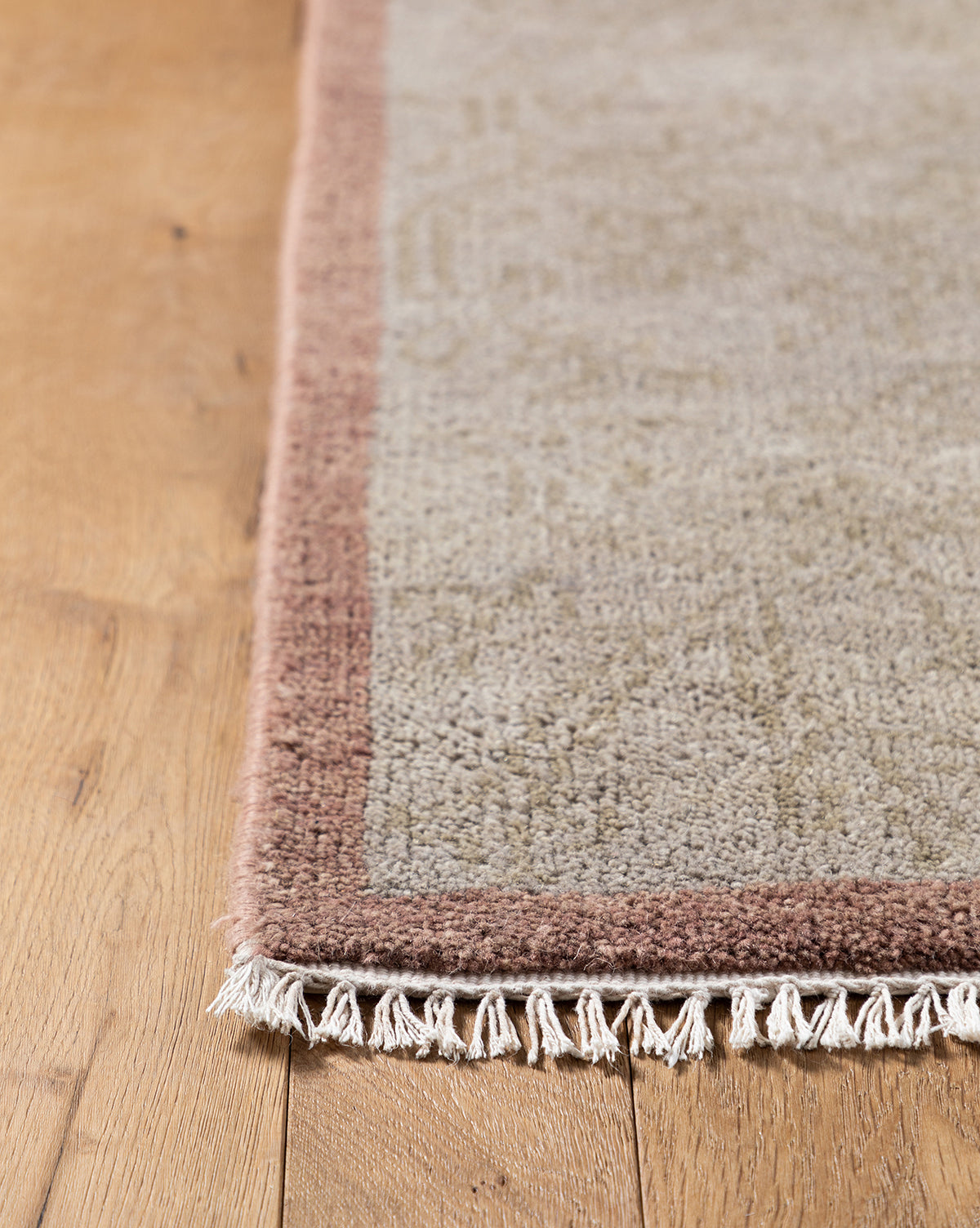 Close-up of the corner of the Suzani Hand-Knotted Wool Rug in light gray with a muted pink border and white fringe, displayed on a wooden floor, by Vendixs
