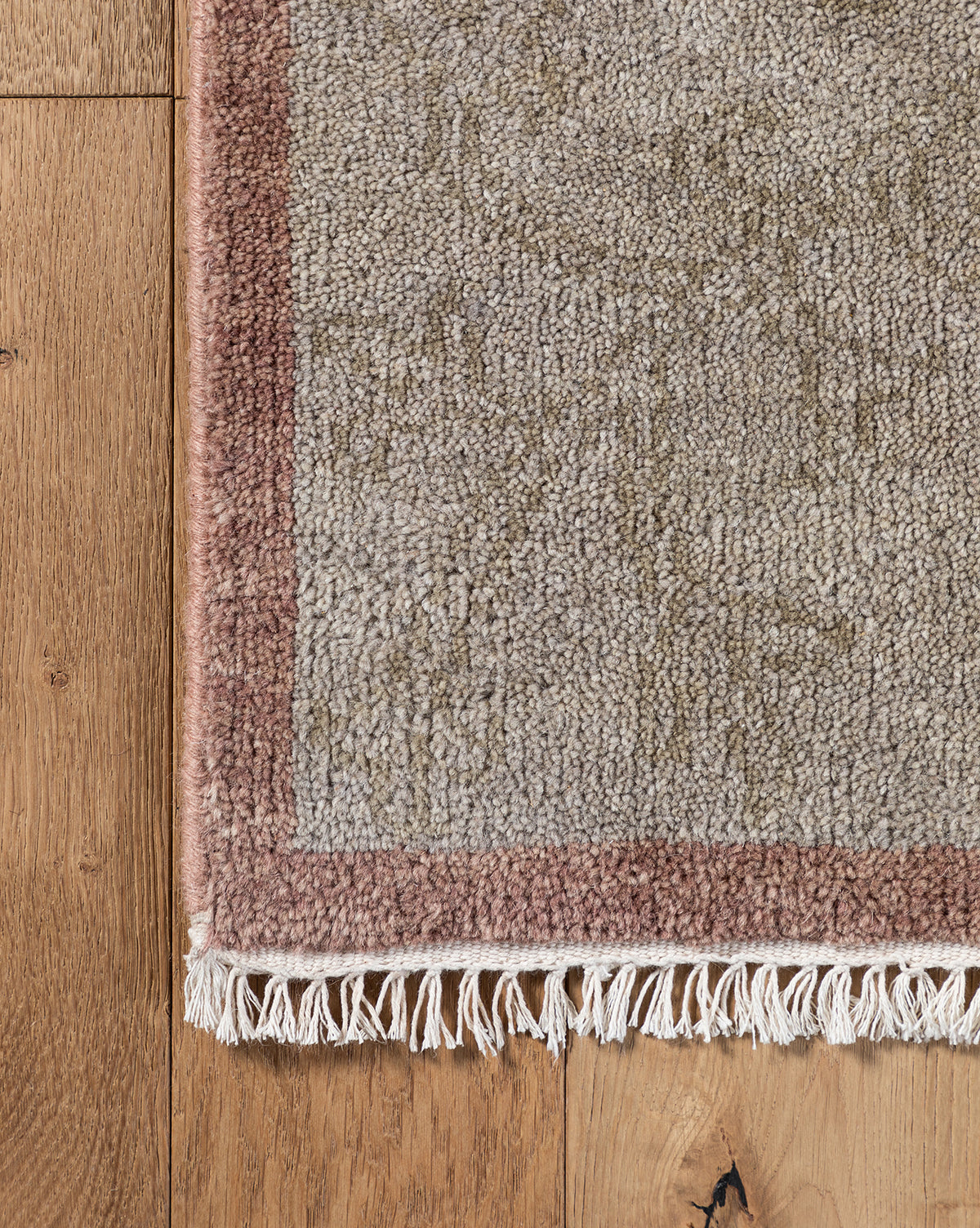 Close-up of the Suzani Hand-Knotted Wool Rug showing a pinkish-brown border, beige center, and white fringe on a wooden floor with visible grain and knots, by Vendixs