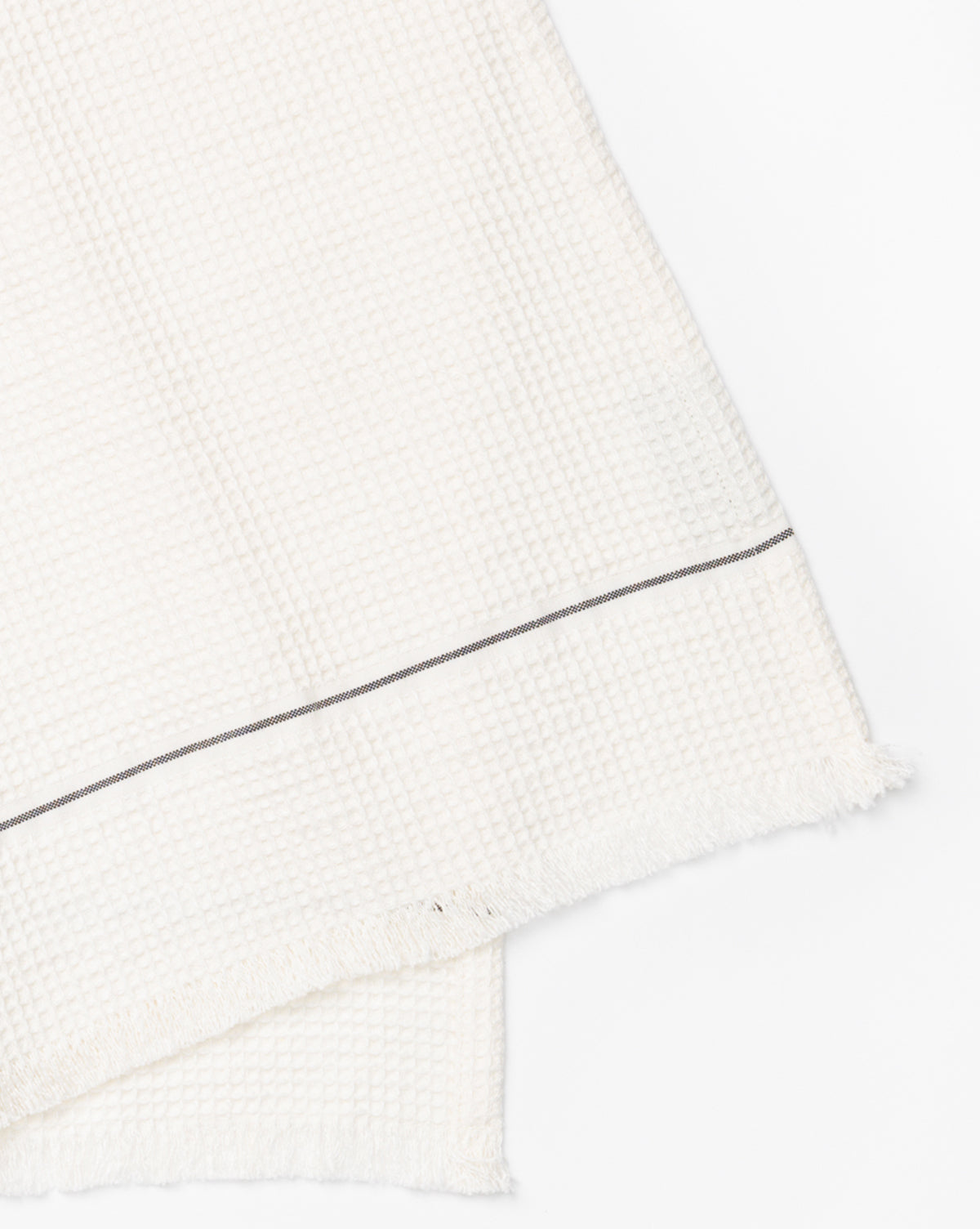 A close-up of the Waffle Hand Towel, made from a linen-cotton blend in a waffle texture with a thin dark stripe and frayed edges, shown on a white background—ideal for adding style to your kitchen decor, by McGee & Co.