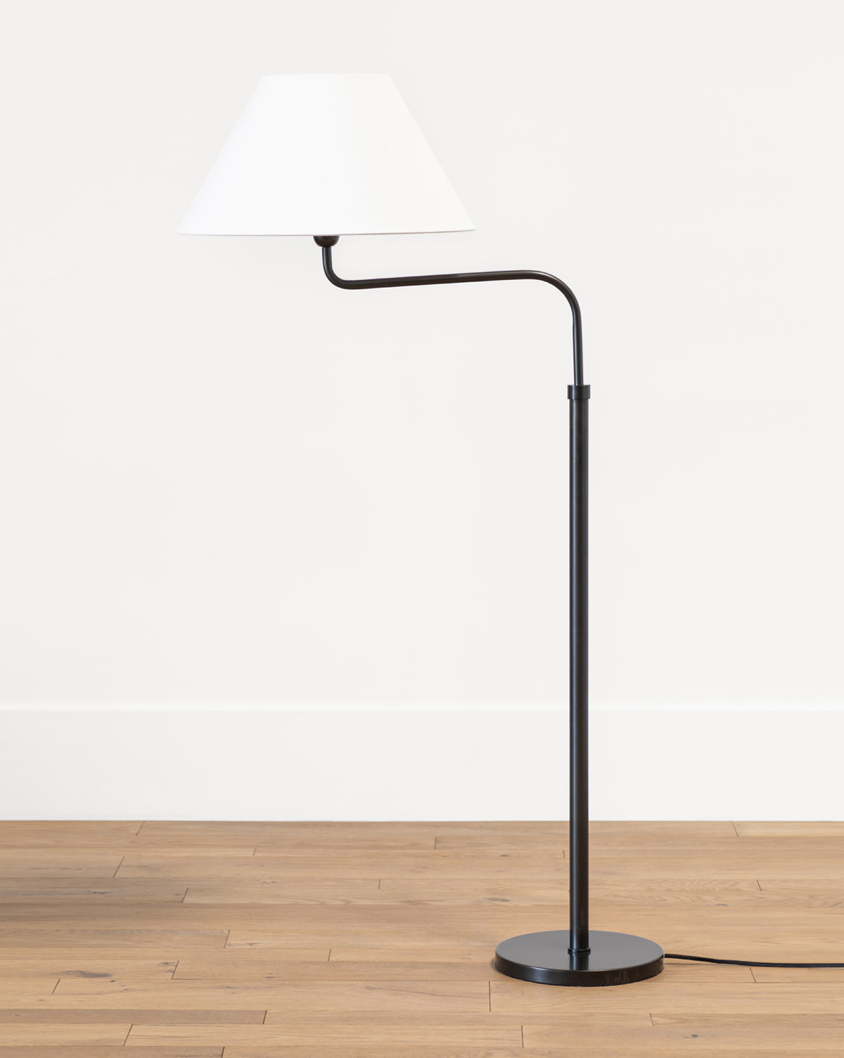 The Makerspalm Wainwright Swoop Floor Lamp features a modern black and brass slim, angled stand with a white conical shade, shown on a wooden floor against a plain white wall. Vendixs