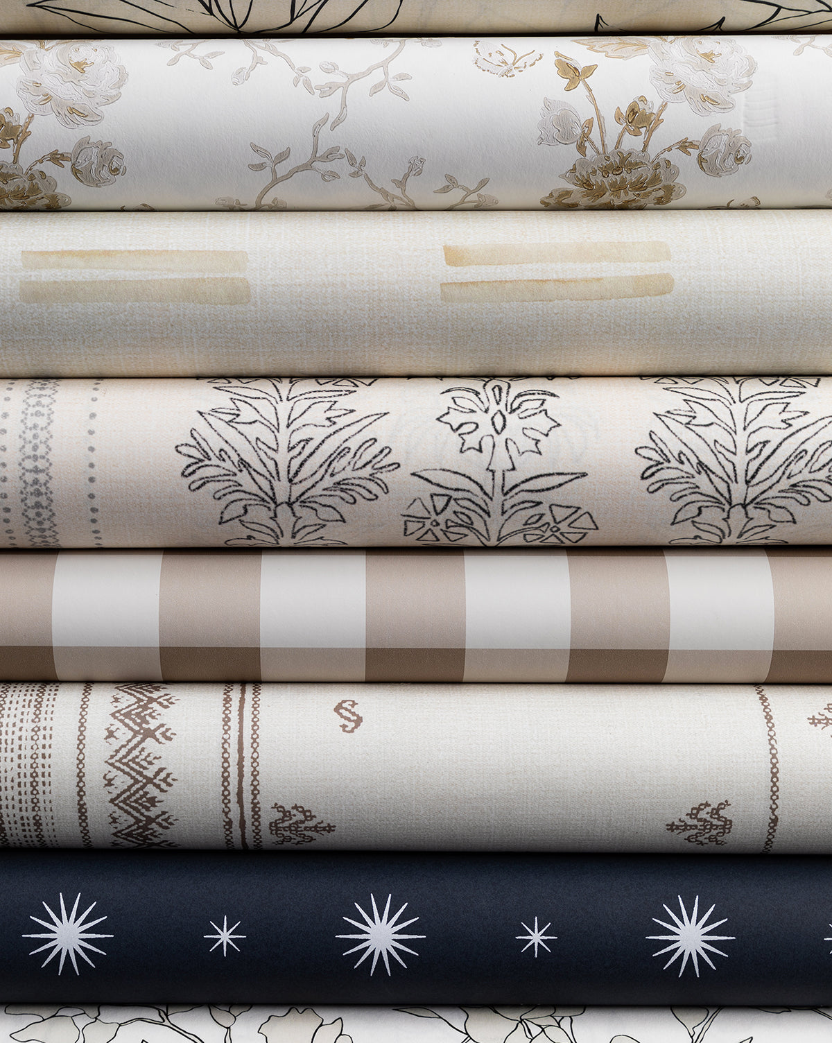A stack of neutral-toned fabrics with florals, stripes, geometrics, and stars—echoing the Demi Floral Stripe Wallpaper by York Wallcoverings—arranged in layers on premium matte paper by Vendixs