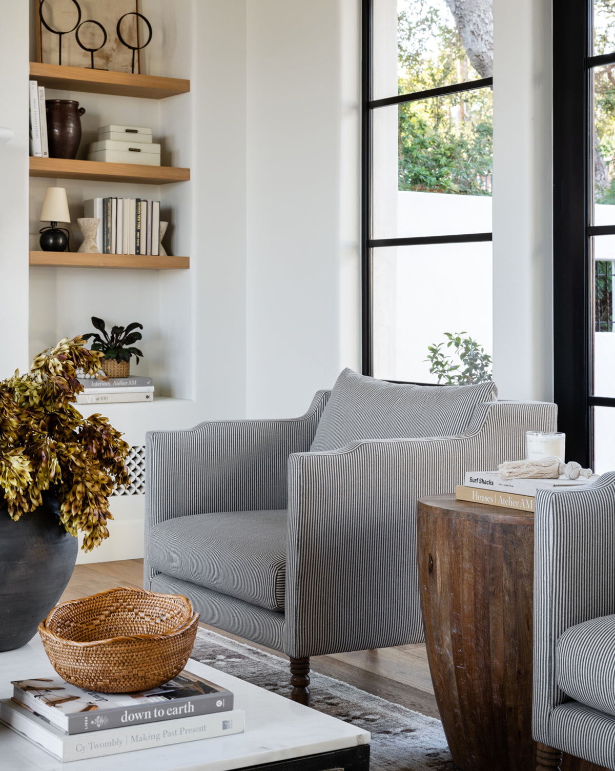 A modern living room with two Gemma Lounge Chairs in premium fabric, a round wooden side table, a woven basket, and a black vase with dried leaves. Built-in shelves with books and decor are in the background next to tall windows, by McGee & Co.