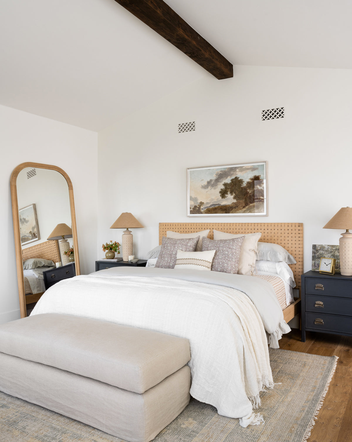 A cozy bedroom features a wooden bed, light bedding, neutral pillows, two black nightstands with lamps, a floor mirror, beige bench, and landscape painting above. The Roslin Hand-Knotted Wool Rug adds warmth alongside the exposed ceiling beam and natural light, by Vendixs