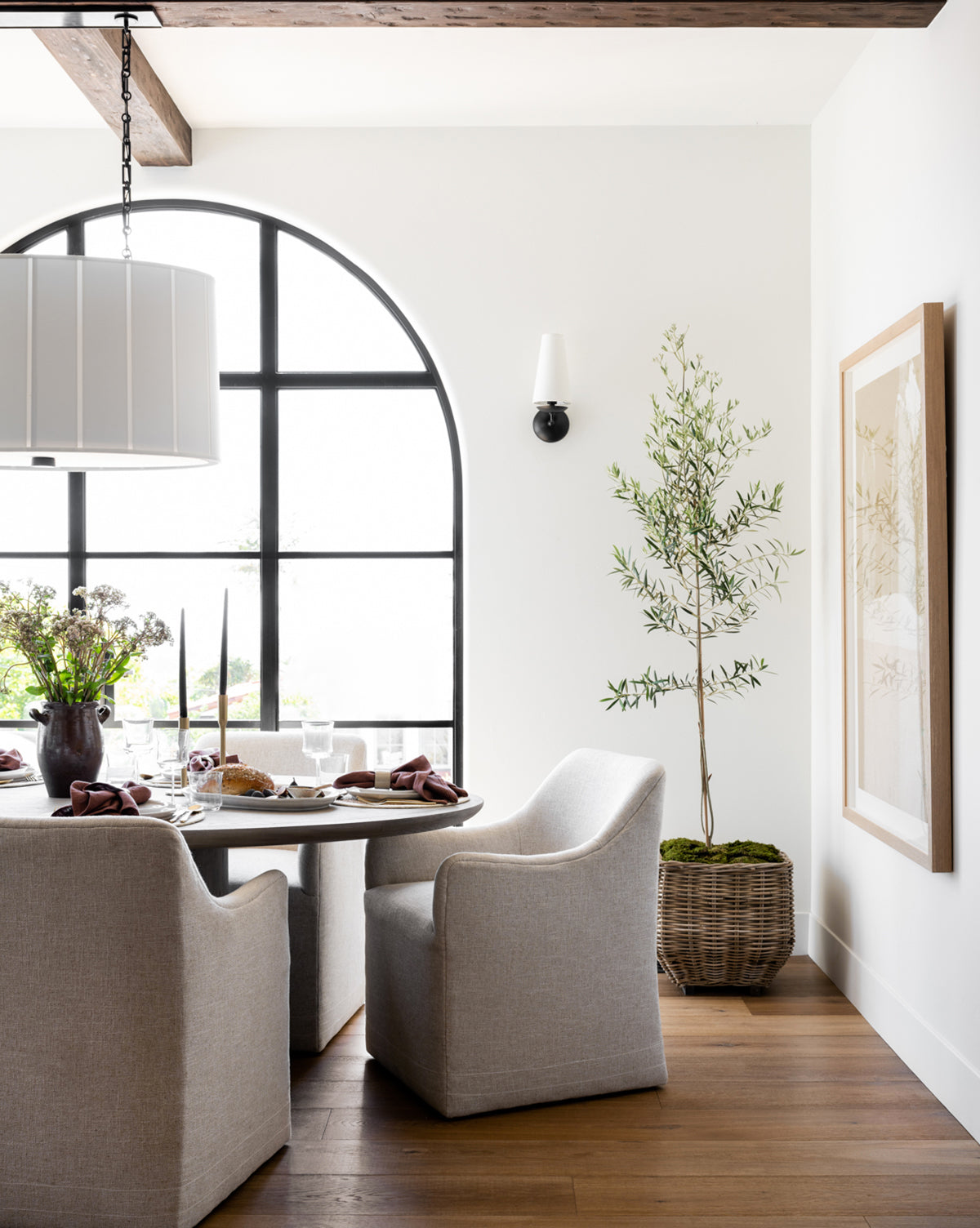Bright dining area with a round table and four Elton Dining Chairs in light linen, large arched window, potted plant, framed wall art, wood flooring; table is set with plates and a floral centerpiece—ideal for stylish dining by Vendixs