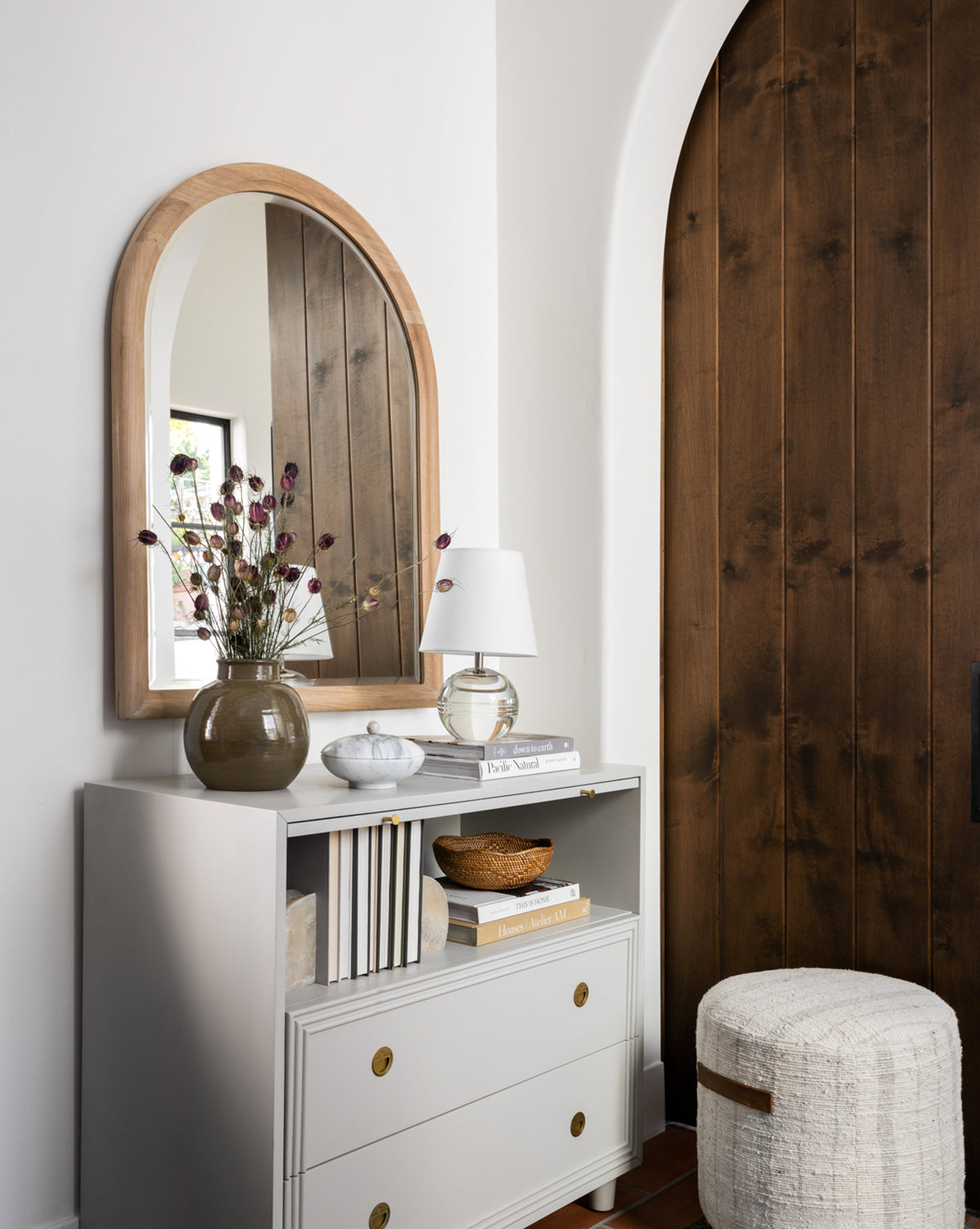 A white dresser with gold handles holds books, a small white lamp, and a brown vase with dried flowers. Above it hangs the Reta Arched Wall Mirror reflecting a window. Nearby are a wooden arched door and a textured round ottoman by Vendixs