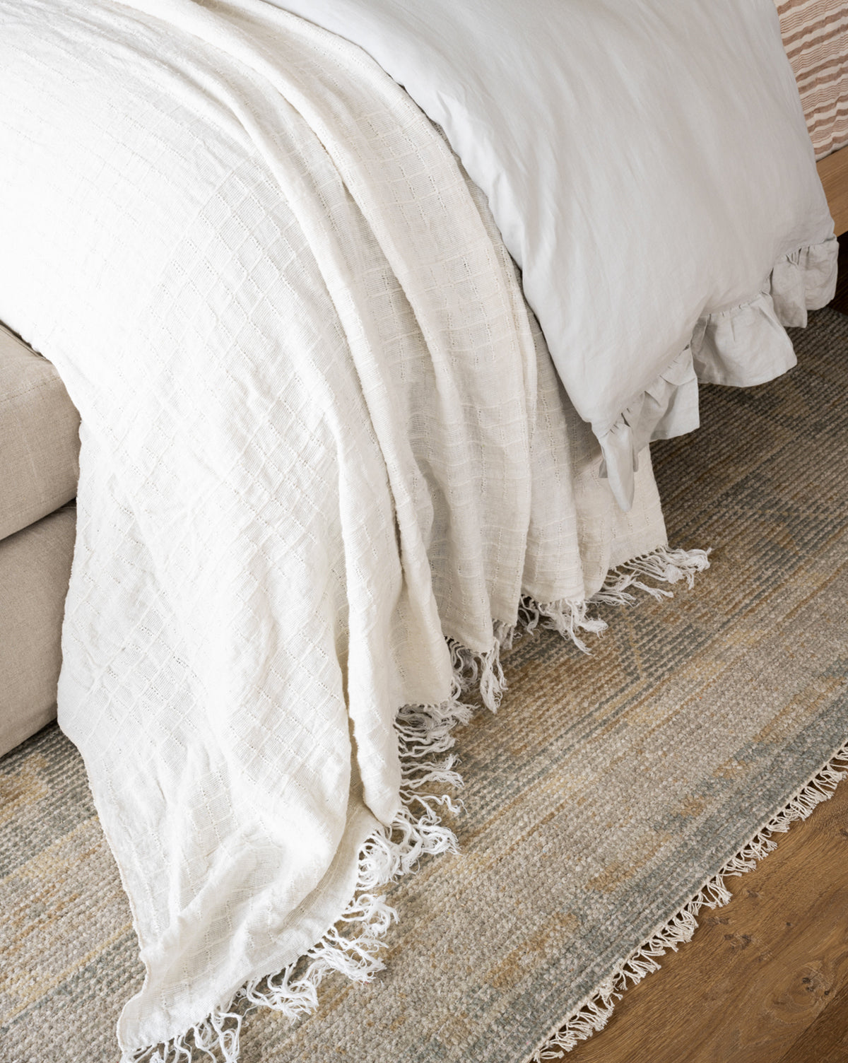 A close-up shows a neatly made bed with a white textured blanket and fringed ends cascading onto the Roslin Hand-Knotted Wool Rug, featuring neutral tones and a subtle geometric pattern on the wooden floor, by Vendixs