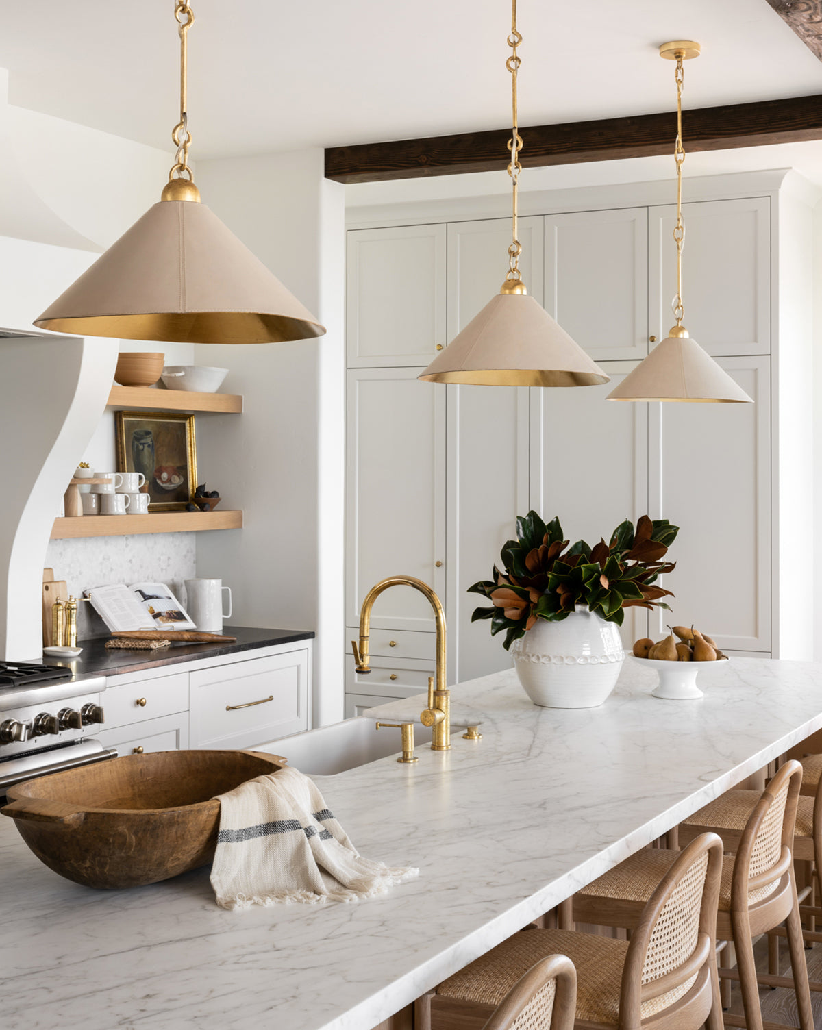 A bright kitchen features white cabinets, marble countertops, gold fixtures, beige pendant lights, woven chairs, a wooden bowl with a Fringed Striped Tea Towel, and a vase of leafy branches on the large island, by Vendixs