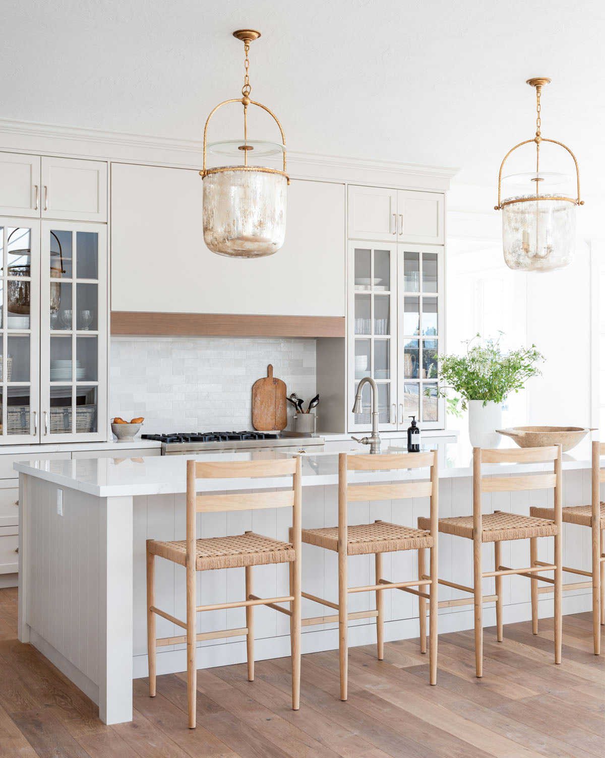 A bright, modern kitchen with white cabinets, a large island with a white countertop, three Eloise Woven Counter Stools, glass pendant lights, and a vase with greenery. Light wood floors complete the airy look, by McGee & Co.