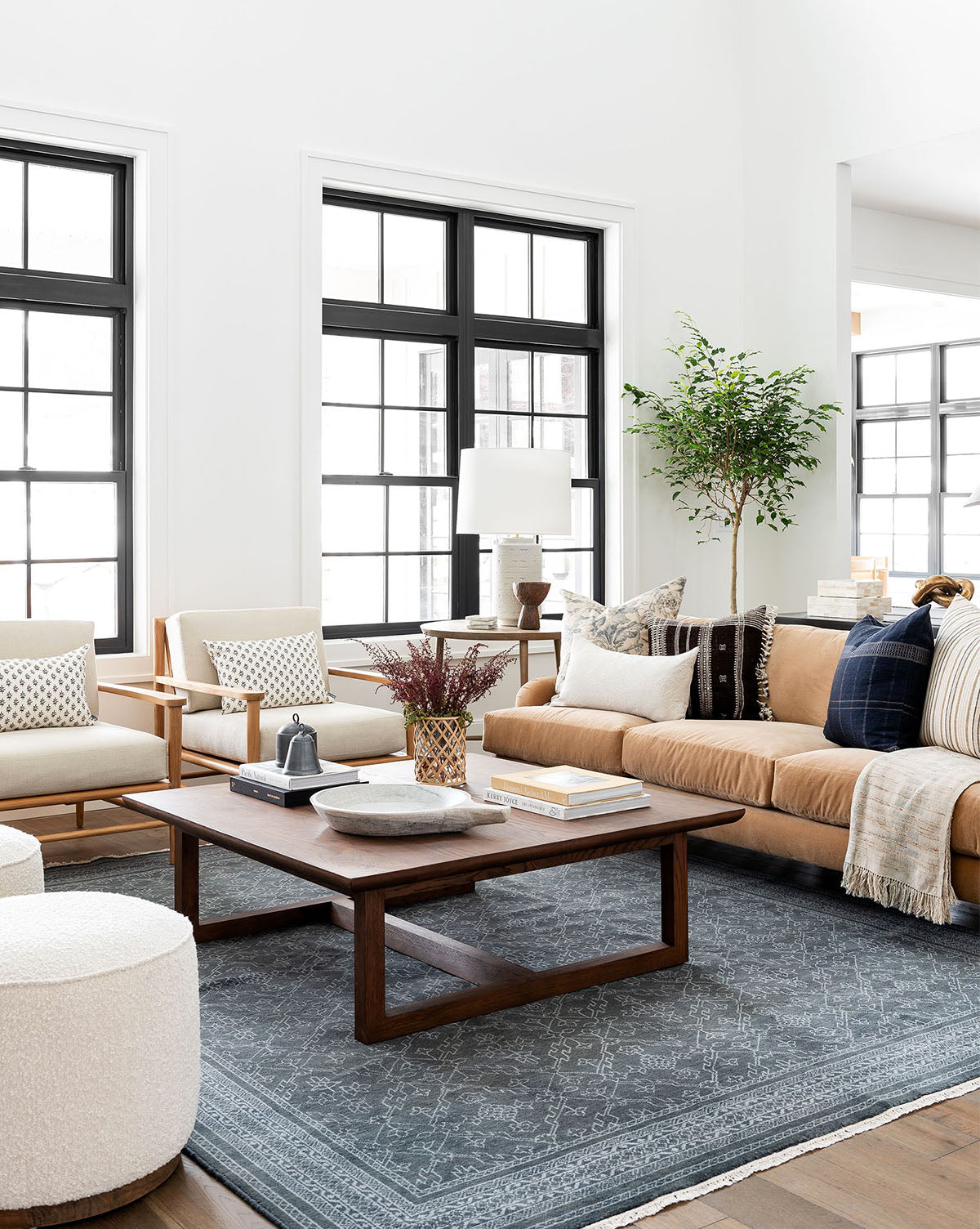 A bright, modern living room with large black-framed windows, a tan sofa, two beige armchairs, the Herrin Coffee Table, a gray patterned rug, decorative pillows, and a potted plant in the corner, by Vendixs