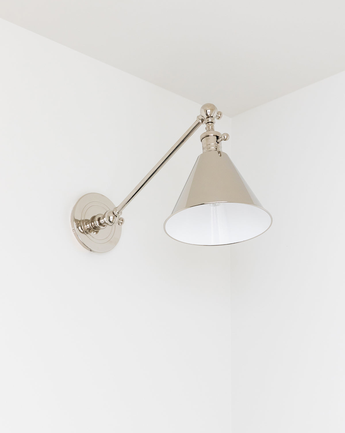 The Boston Single Arm Library Light by Visual Comfort & Co. Signature, with its modern metallic design and conical shade, is mounted on a white wall in the corner, complementing the room’s minimalist decor. Vendixs