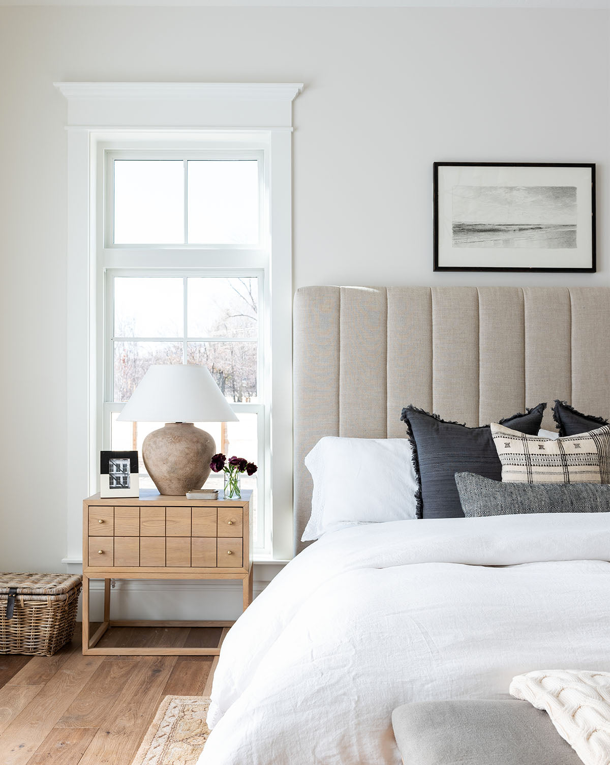 Bright, modern bedroom with a beige Hoffman Bed Tall Headboard in customizable fabric, white bedding, neutral pillows, wooden nightstand with lamp and framed photo, flowers, large sunlit window, and wicker basket on the floor, by McGee & Co.