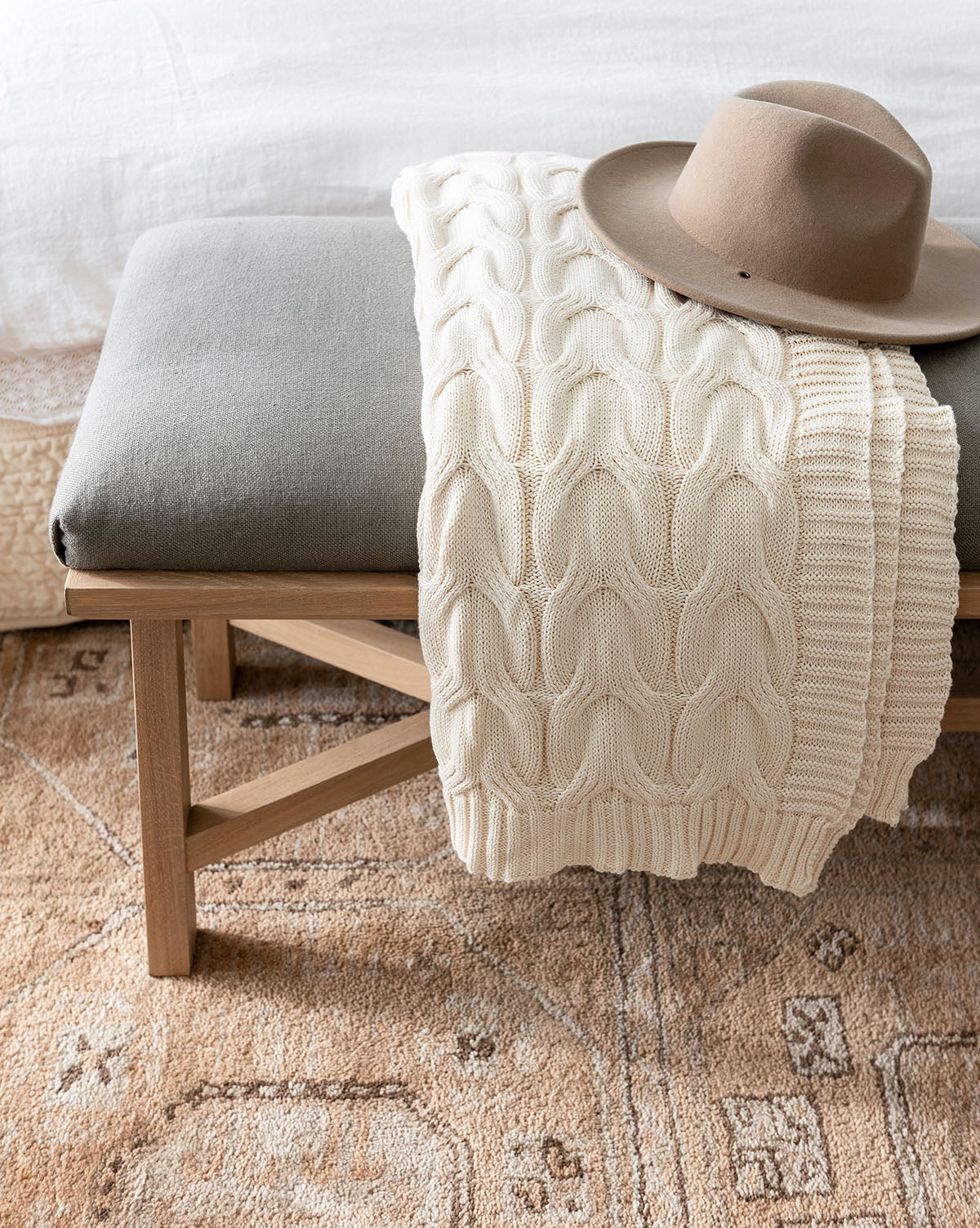 A beige knit blanket and a brown wide-brimmed hat rest on a cushioned wooden bench at the foot of a bed, atop the Ellington Fawn Hand-Tufted Wool Rug, by Vendixs