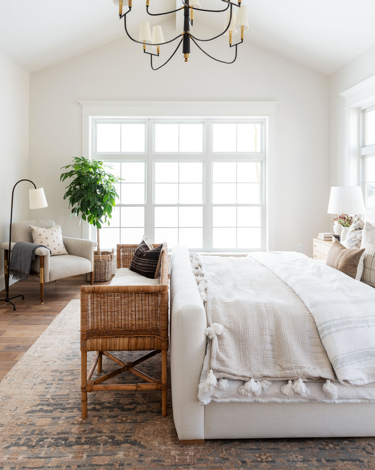 Bright, airy bedroom with large windows, a cozy armchair, potted plant, Wilshire Blue Hand-Tufted Wool Rug beside a woven bench at the bed’s foot. Light bedding, neutral decor, wood floor, and a chandelier complete the inviting space, by Vendixs