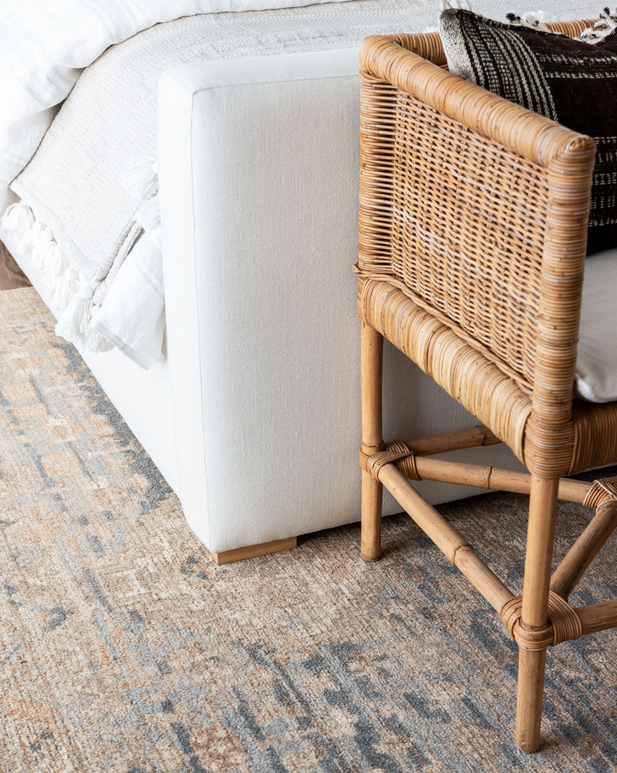 A close-up of a cozy bedroom corner featuring a white upholstered bed, a woven rattan chair with patterned pillows, and the Wilshire Blue Hand-Tufted Wool Rug, by Vendixs