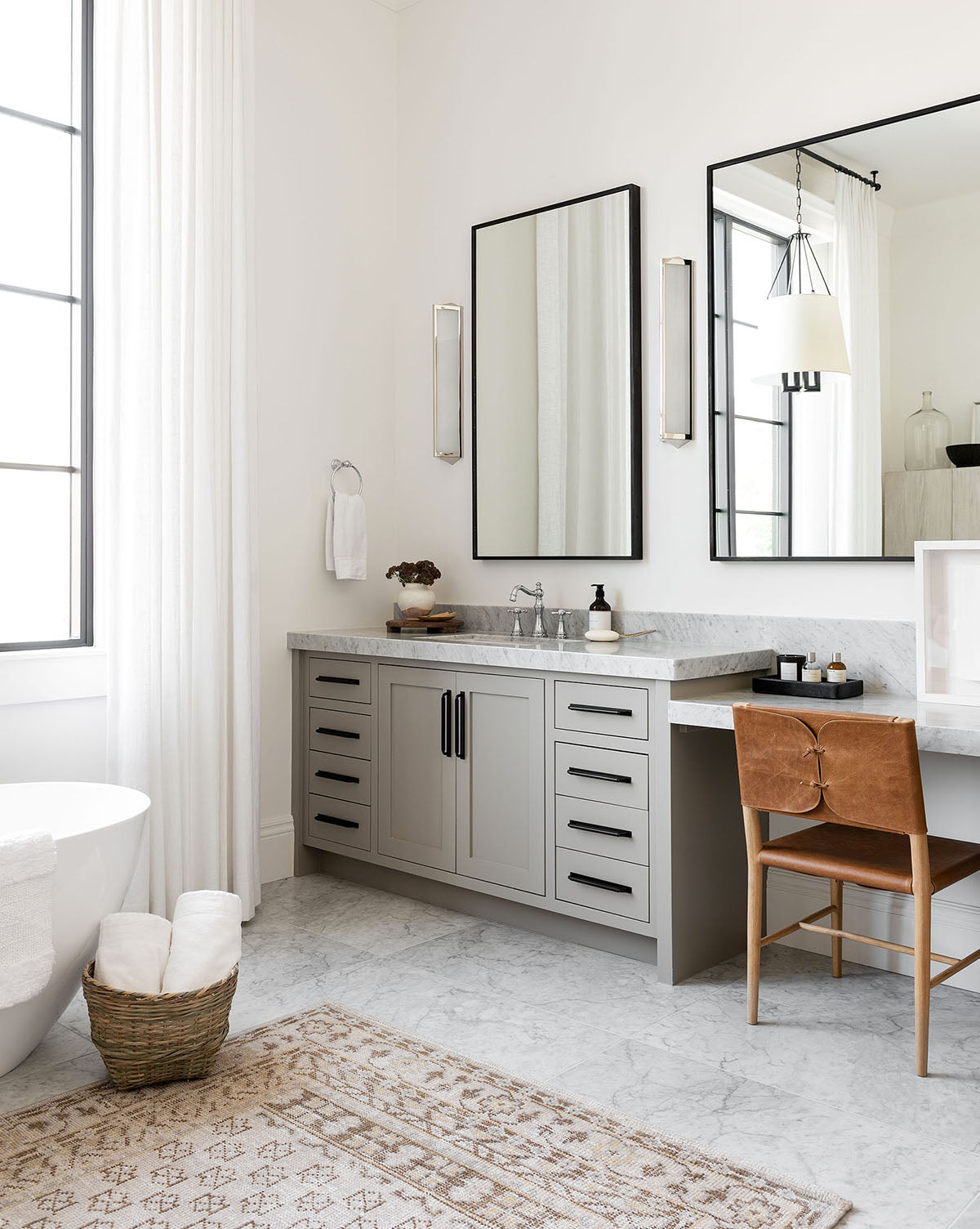 A modern bathroom features a freestanding bathtub, light gray double-sink vanity, large mirrors, brown leather chair, marble floors, and the Anya Neutral Hand-Knotted Wool Rug paired with a basket of rolled towels for added comfort, by Vendixs