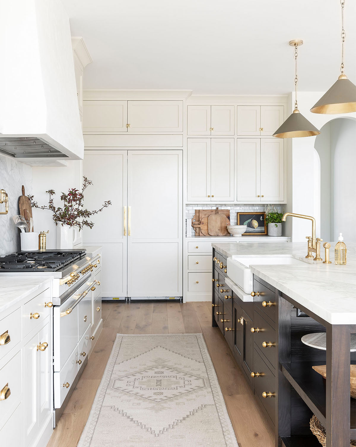 Bright, elegant kitchen with white cabinets, gold hardware, marble countertops, a farmhouse sink, pendant lights, and the Pembroke Hand-Knotted Wool Rug on light wood floors. Decorative plants and dishes adorn the counters and shelves, by Vendixs