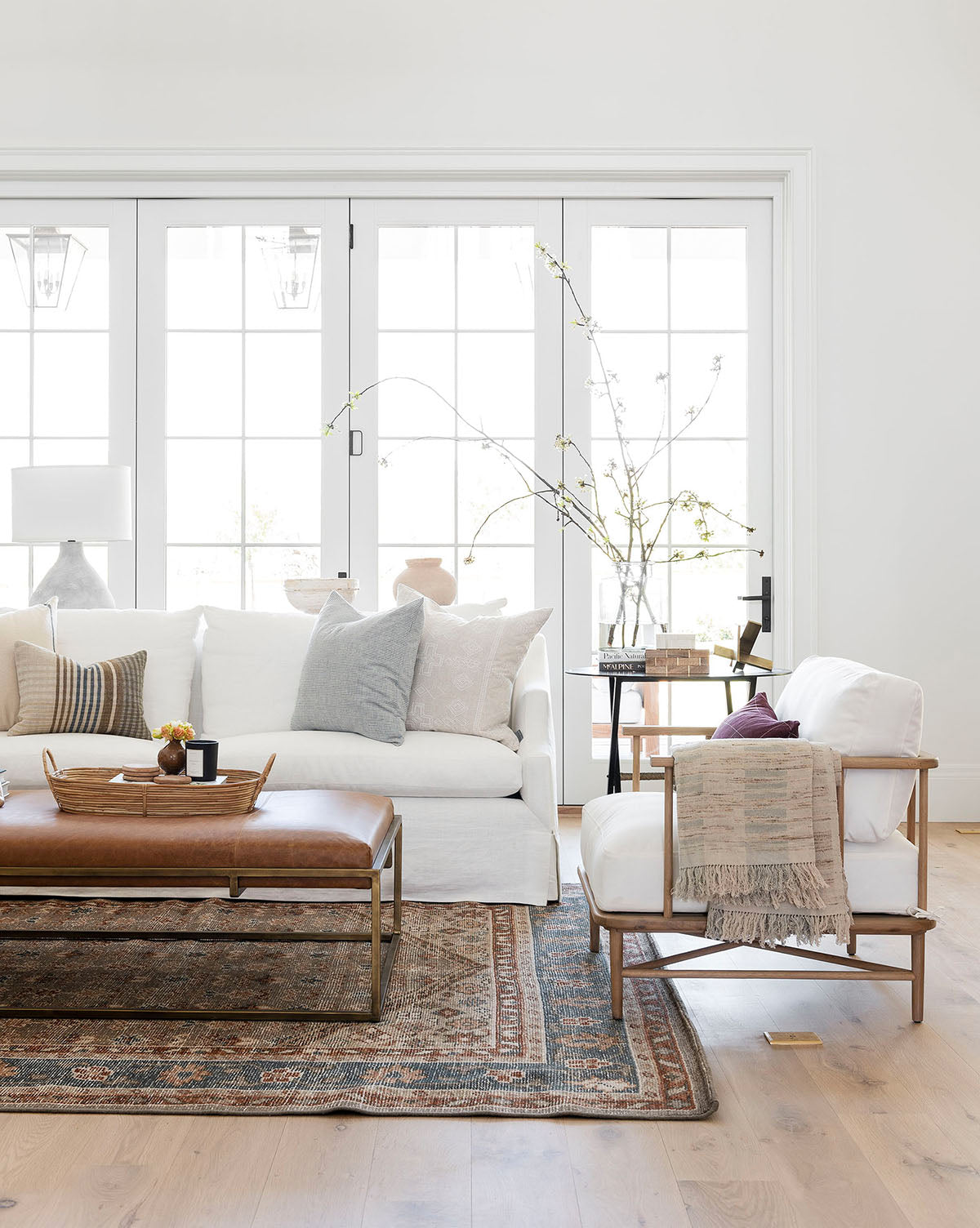 Bright, modern living room features a white sofa, armchair, patterned rug, soft pillows, decorative vases, large windows with natural light, and the Harlow Leather Coffee Table with brass accents, by McGee & Co.
