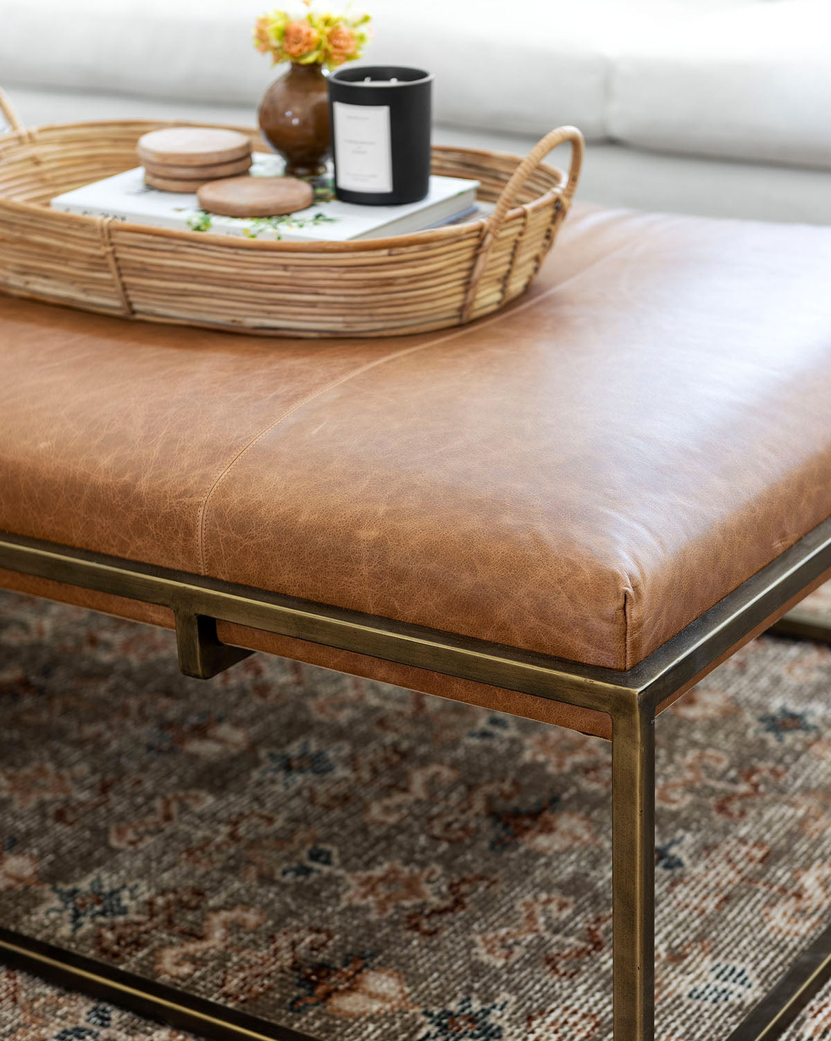 The Harlow Leather Coffee Table features an aniline-dyed rectangular leather top with a modern brass-finished frame. A woven tray holds a black candle, coasters, and yellow flowers. The ottoman rests on a patterned rug, by Vendixs