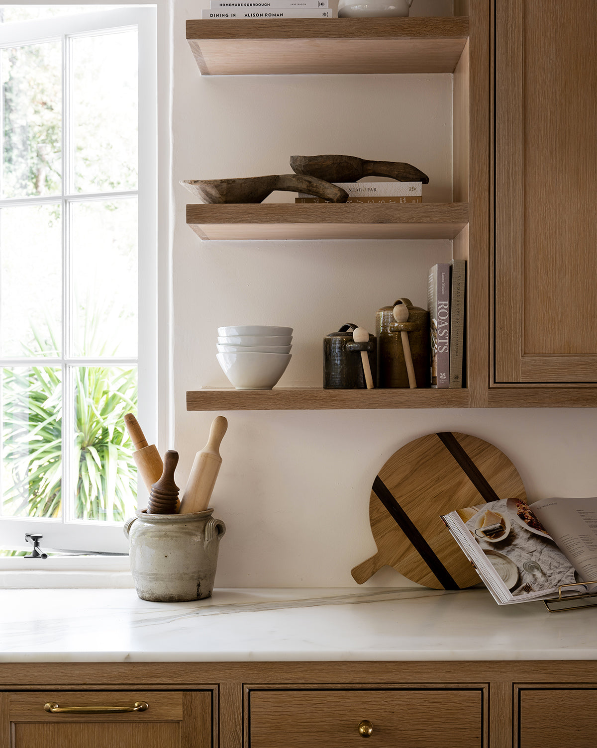 A bright kitchen corner with wooden shelves displays bowls, books, and utensils. Sunlight highlights a marble counter set with a crock of rolling pins, cutting boards like the Round Oak Bread Board, a pepper mill, and an open cookbook by Vendixs