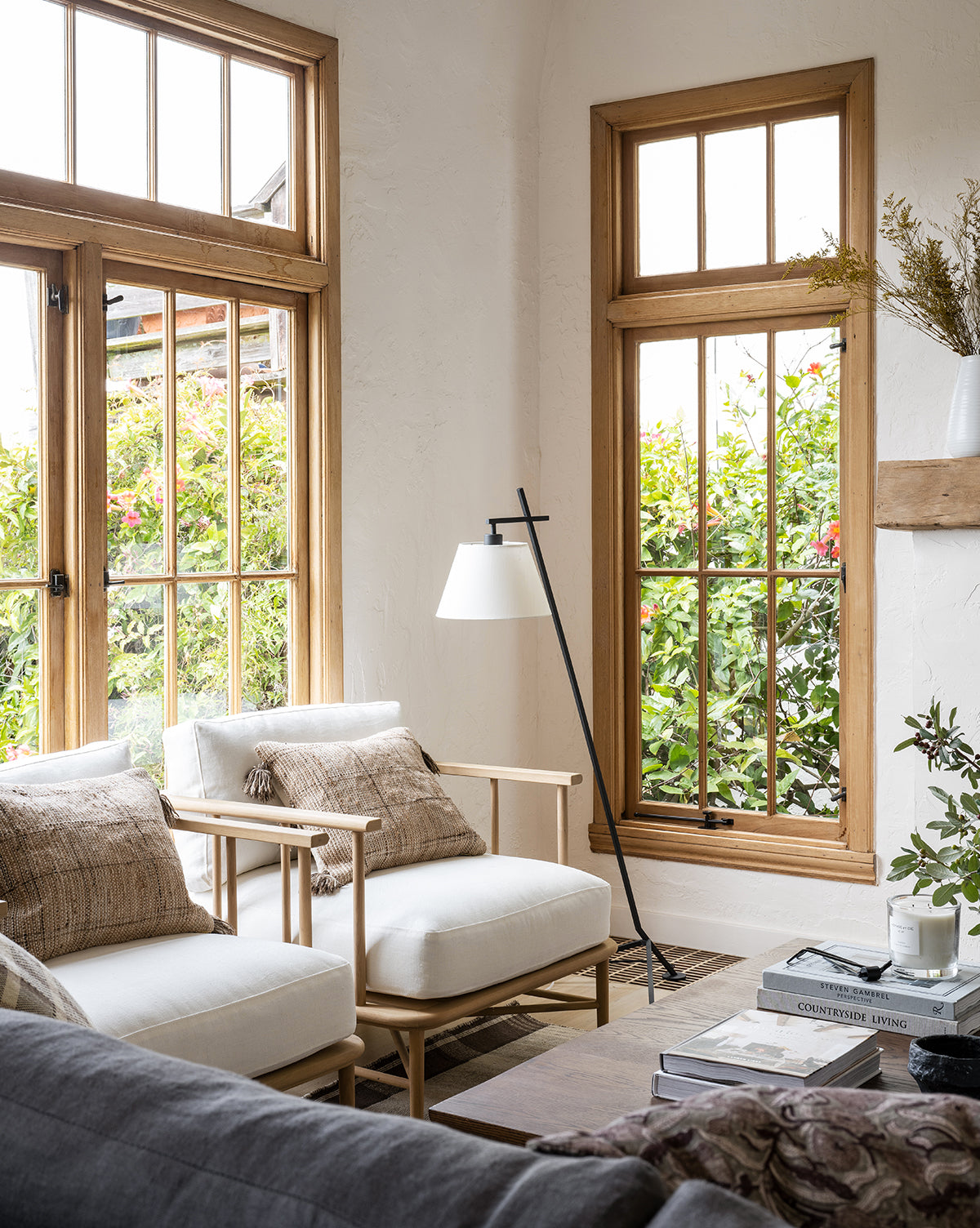 Bright living room with large windows, wooden frames, an Alma Lounge Chair with plush cushions, two cushioned chairs with textured pillows, a modern floor lamp, coffee table stacked with books, and greenery outside. The space feels cozy and inviting by McGee & Co.