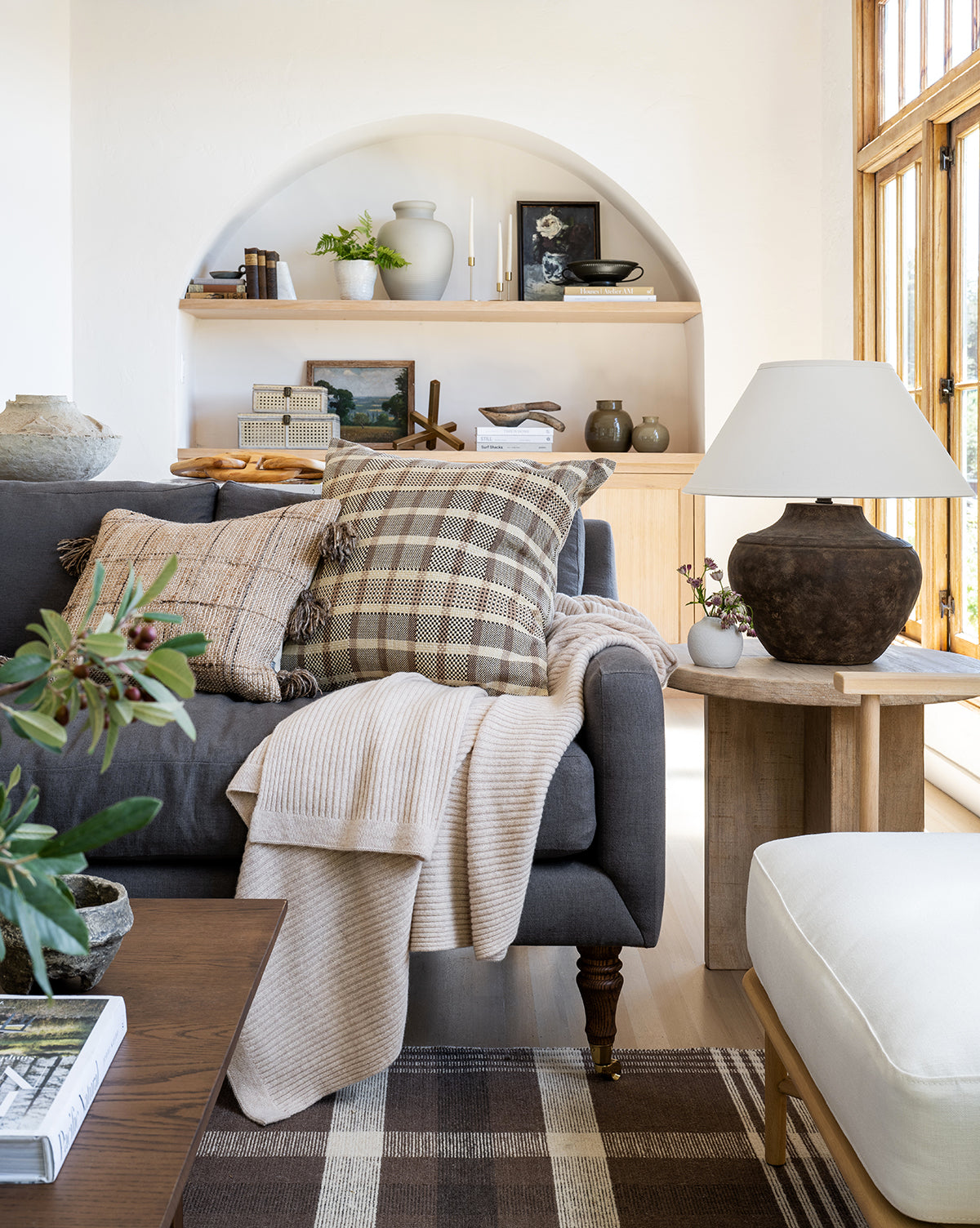 A cozy living room with a Barden Sofa 84 featuring tufted cushions and reeded wood legs, plaid and beige pillows, a cream throw, a rustic side table with a lamp, built-in shelves with books and plants, and sunlight through tall windows by Vendixs