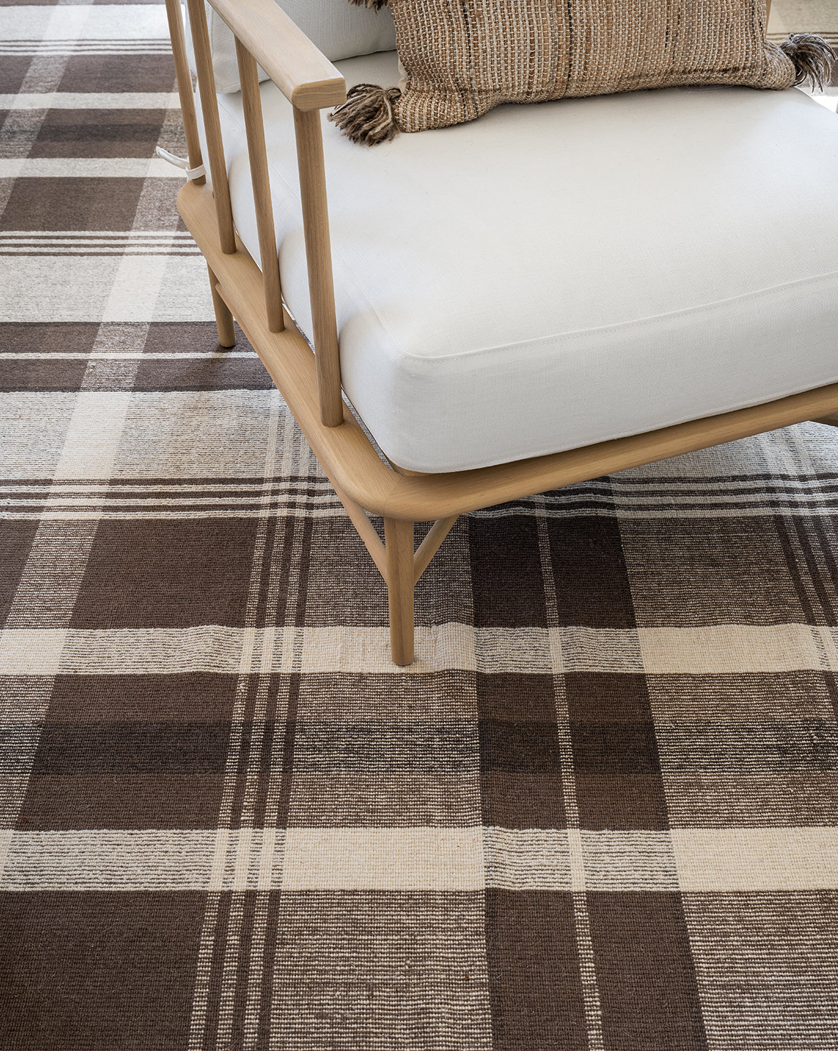 A light wooden armchair with white cushions sits on the Orton Handwoven Wool Rug, which features a brown and beige plaid pattern. A textured striped pillow with tassels is placed on the chair, by Vendixs