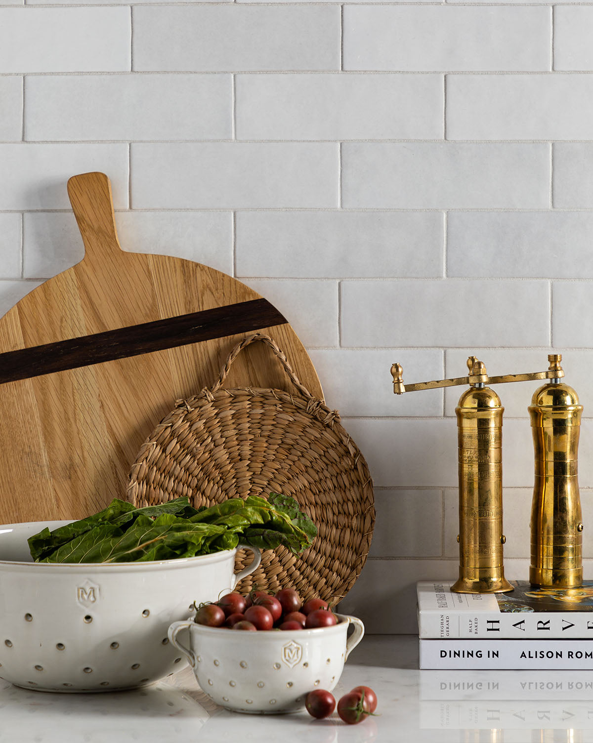 A kitchen counter with two Glazed Colanders of greens and cherry tomatoes, stylish serveware, a wooden cutting board, a woven trivet, brass pepper mills, and cookbooks against white subway tiles for chic decor, by Vendixs