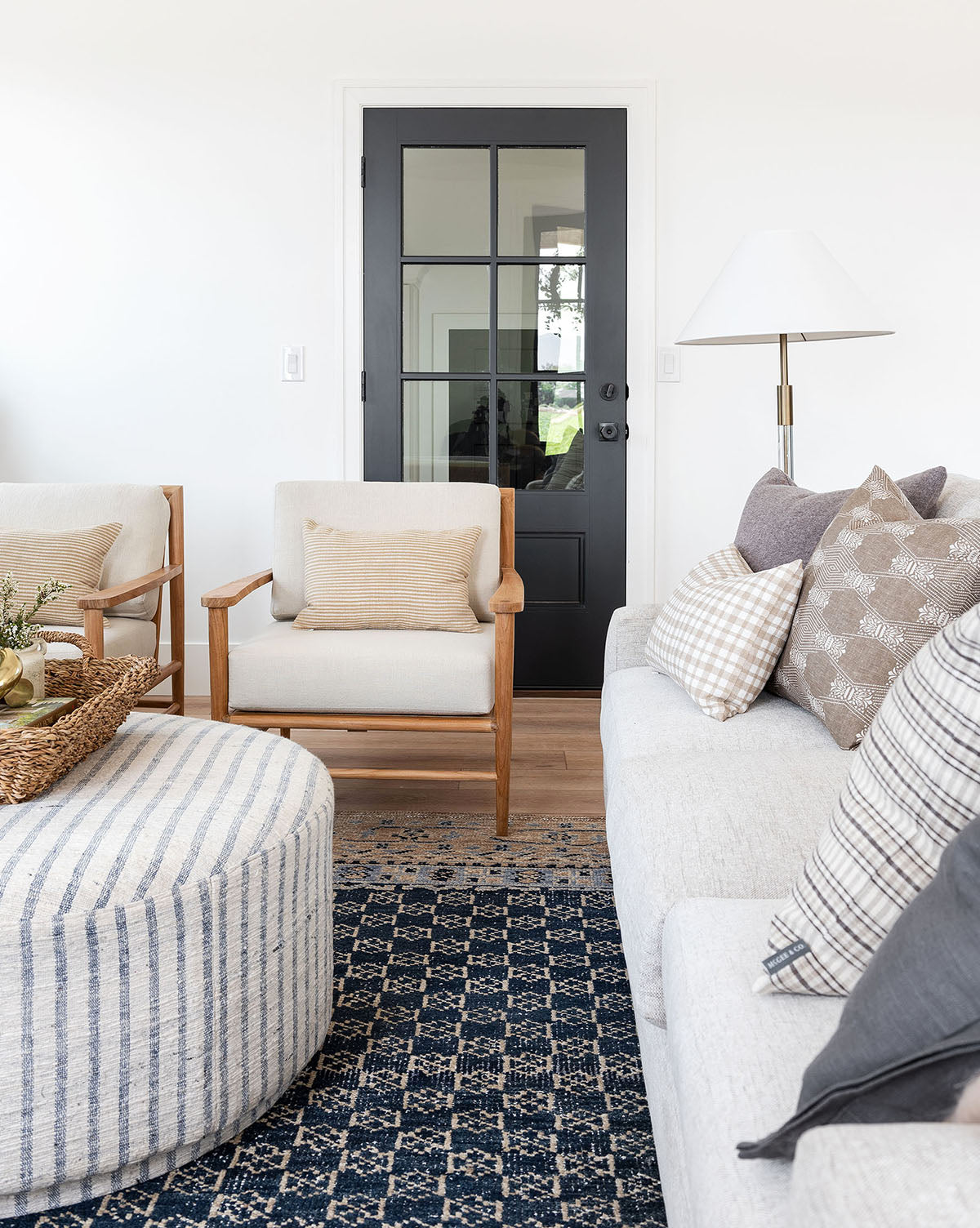 A bright, modern living room featuring a black door, beige armchair, light gray sofa with patterned pillows, striped ottoman, and tall floor lamp is anchored by the Anya Indigo Hand-Knotted Wool Rug for added elegance, by Vendixs
