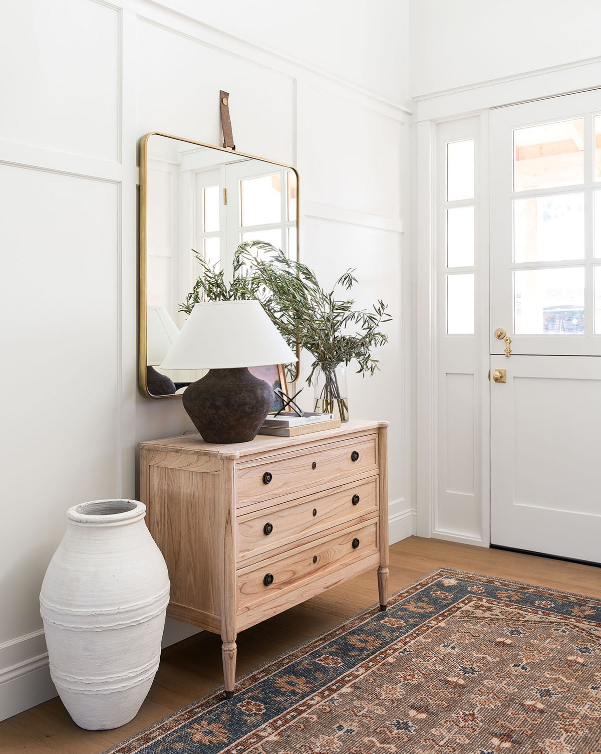 A light wood dresser with brass hardware, a brown lamp, a leafy plant in a vase, and the Laila Rectangle Mirror above sits near a white front door. A tall white ceramic vase and patterned rug complete the stylish entryway by Vendixs