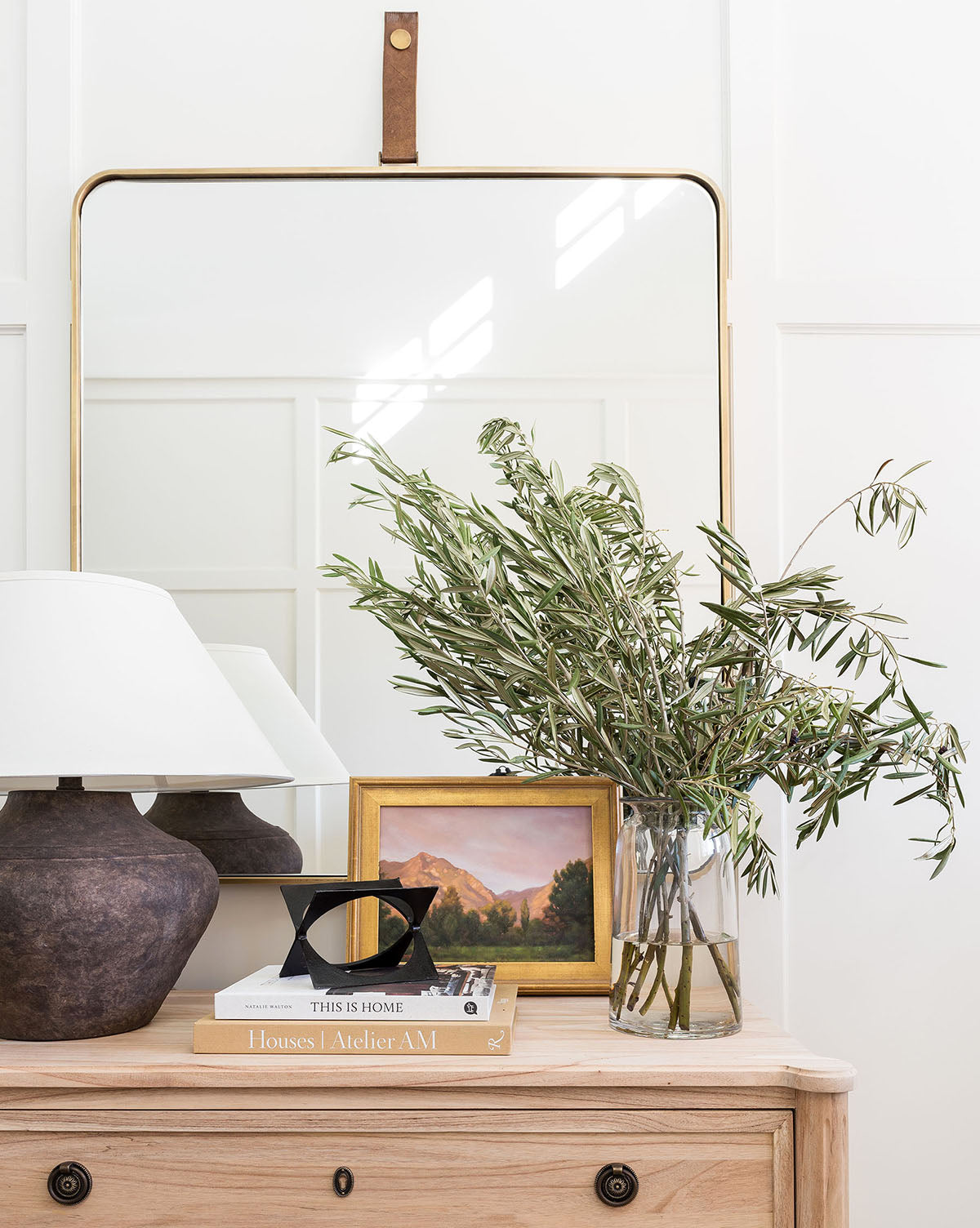 A wooden dresser with brass hardware is styled with a dark lamp, glass vase of leafy branches, small framed landscape painting, decor accents, and two stacked books. The Laila Rectangle Mirror is mounted on the wall behind in a gold frame by Vendixs
