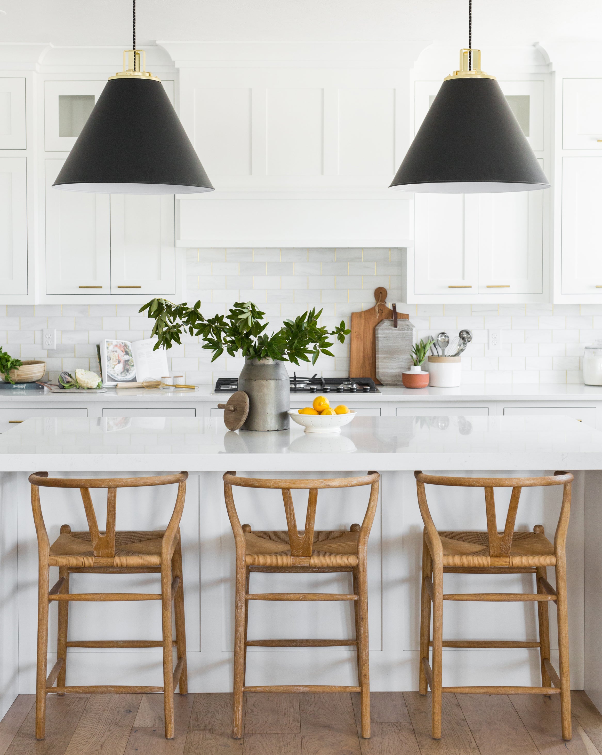 Modern kitchen with white cabinets, a large island, three Gabriel Counter Stools with rope seats and oak frames inspired by Scandinavian Modernism, black pendant lights, plus decorative touches like a bowl of lemons and green leafy branches, by Vendixs