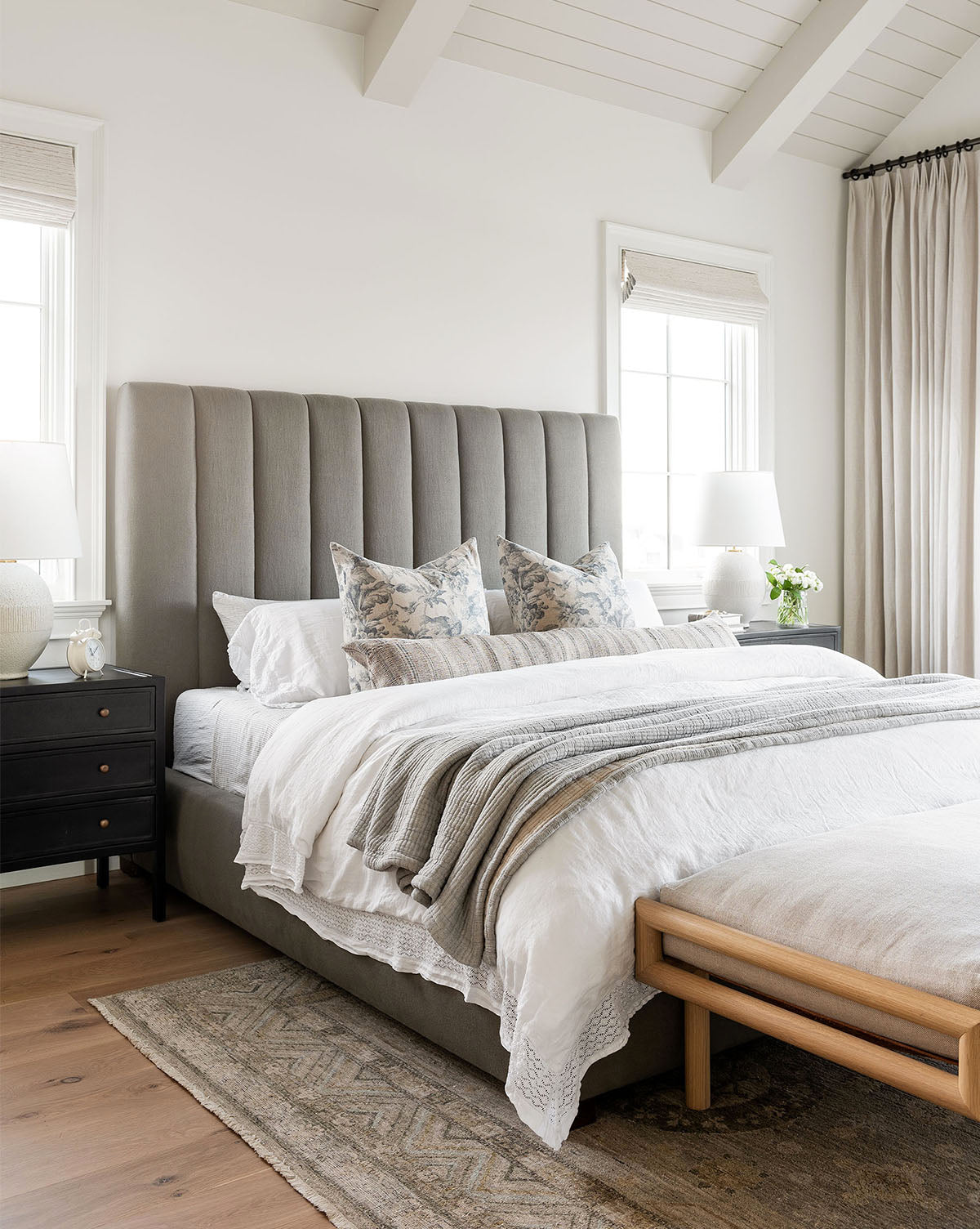A modern bedroom with the Hoffman Bed Tall Headboard in customizable upholstery, white bedding, patterned pillows, and a gray throw. Black nightstands with lamps and a wooden bench finish the look as large windows fill the space with light, by McGee & Co.